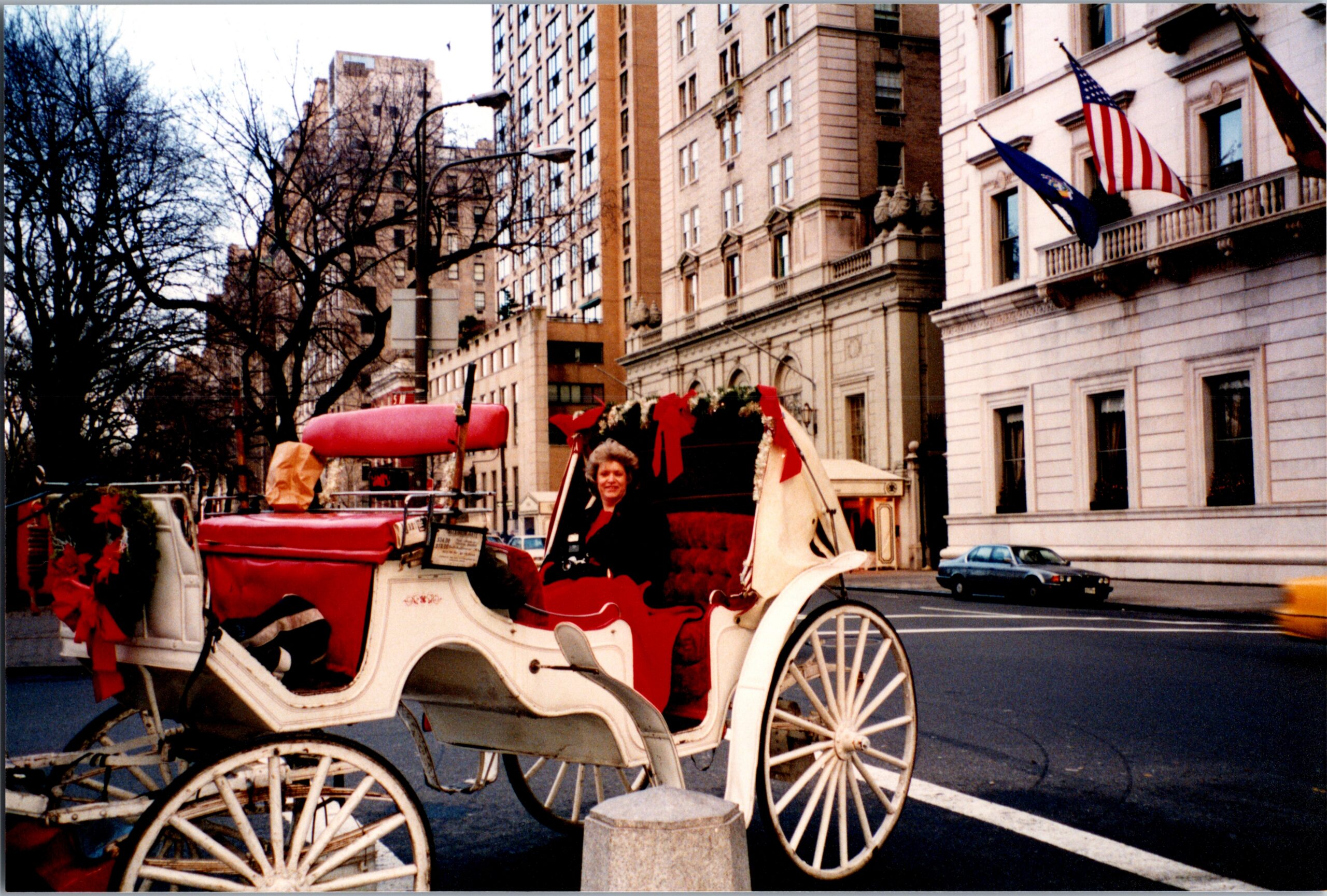 White decorative horse-drawn carriage with red seating on a city street; a woman sits at the reins, with flags flying from a building nearby.