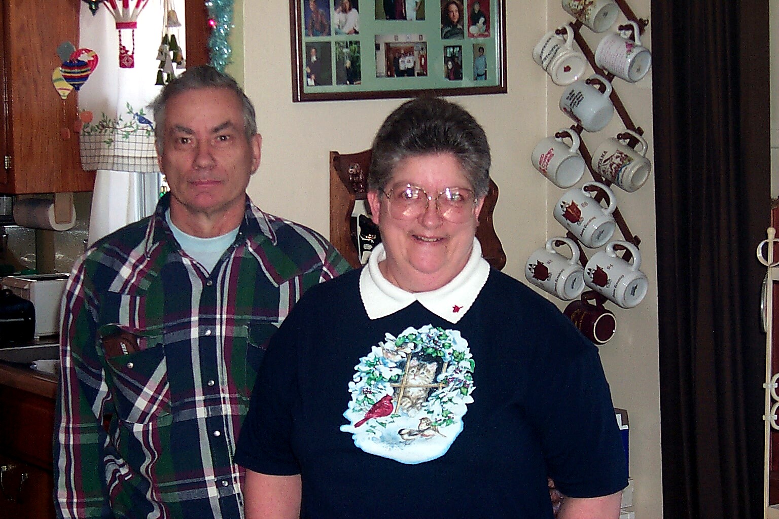 Smiling couple stands in a cozy kitchen decorated for Christmas, with a mug rack visible behind them on the wall.