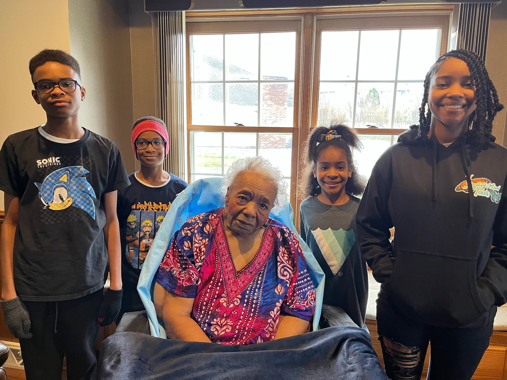 Group portrait of six people indoors by a large window: two boys standing on the left, an elderly woman seated center, a young girl with a ponytail, another girl with braids, and a smiling teen girl on the right.