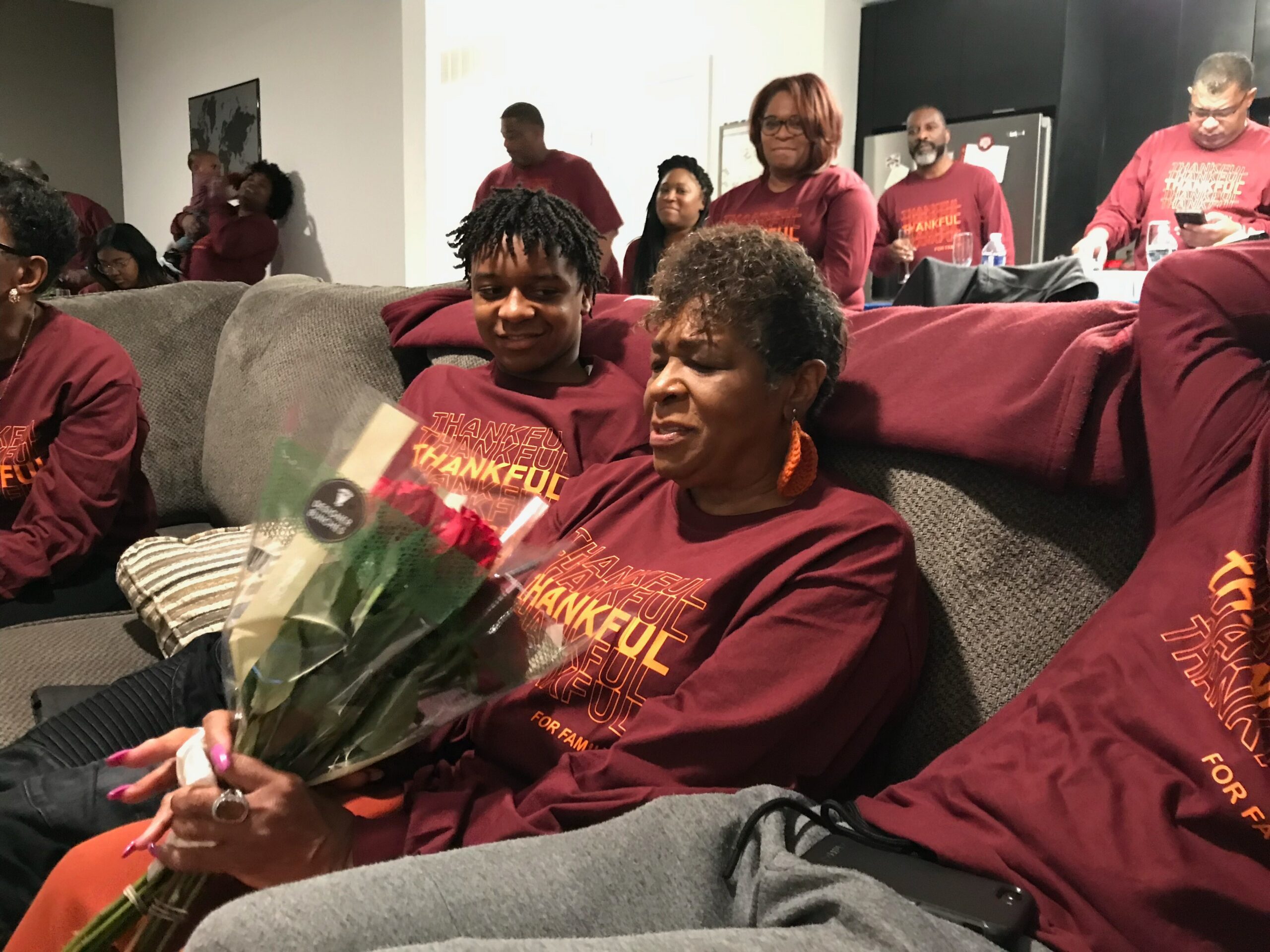 Group of adults in maroon shirts in a living room, with a woman in front holding a bouquet of flowers.