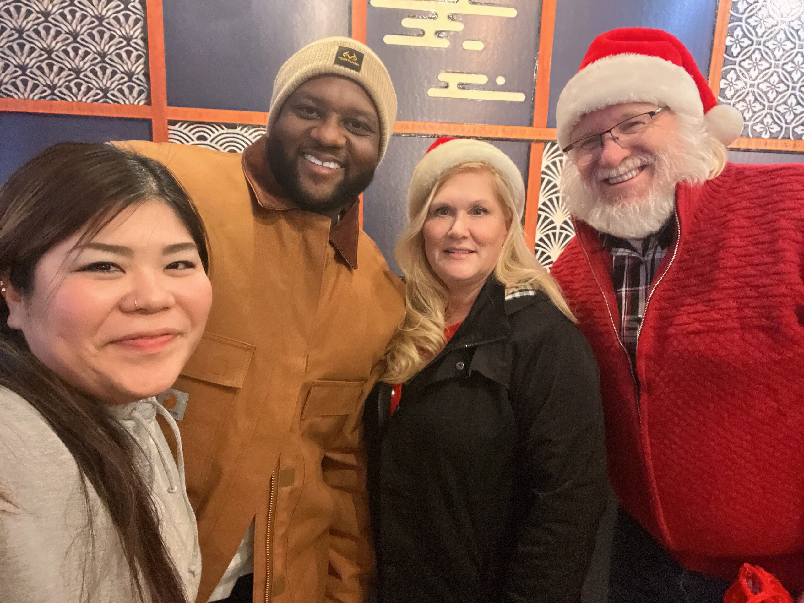 Four people posing for a selfie indoors: woman in a gray hoodie in the foreground, a man in a tan jacket and beige beanie, a blonde woman, and a Santa Claus in red suit smiling together.