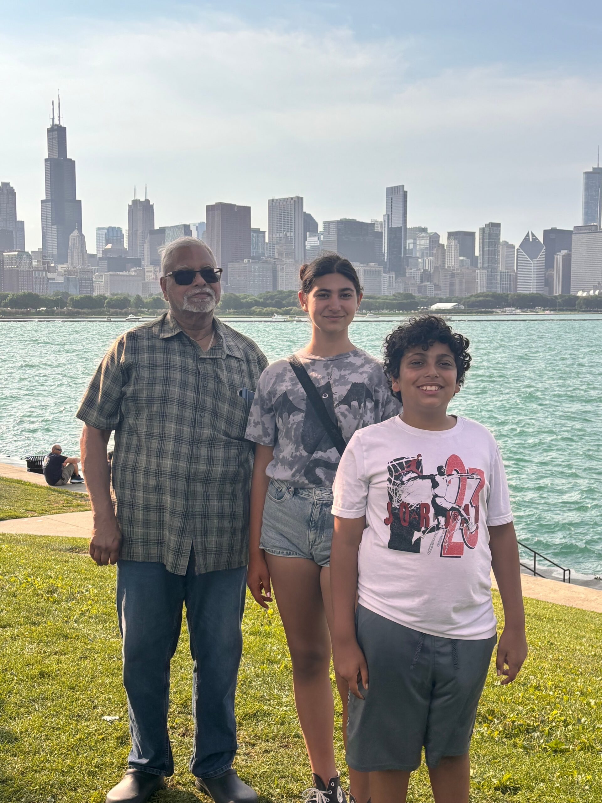 Three people stand together on a grassy lakeside with a city skyline in the background.