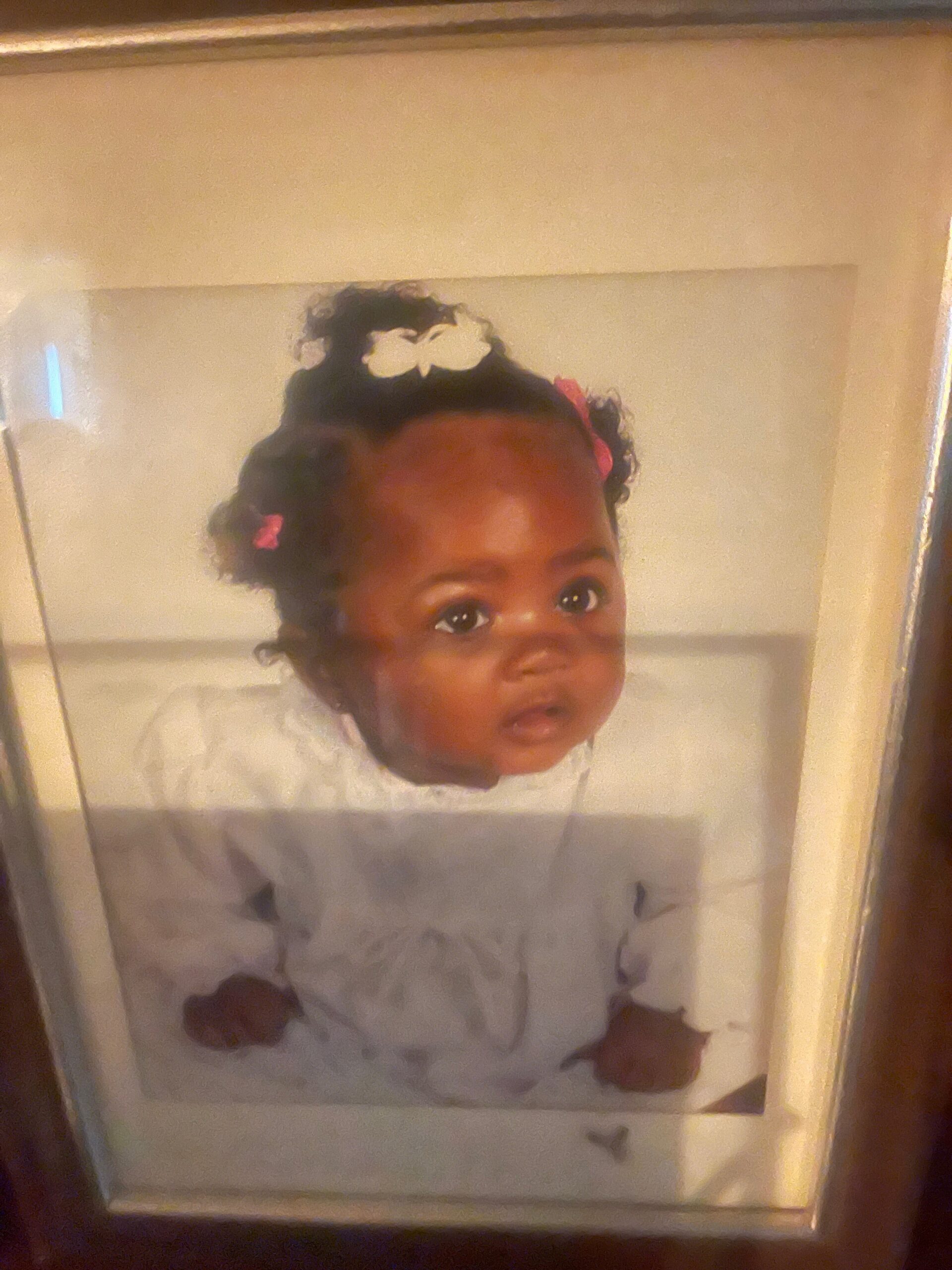 Doll portrait: young girl with dark skin, curly hair, white bow and pink clips, wearing white clothing inside a frame.