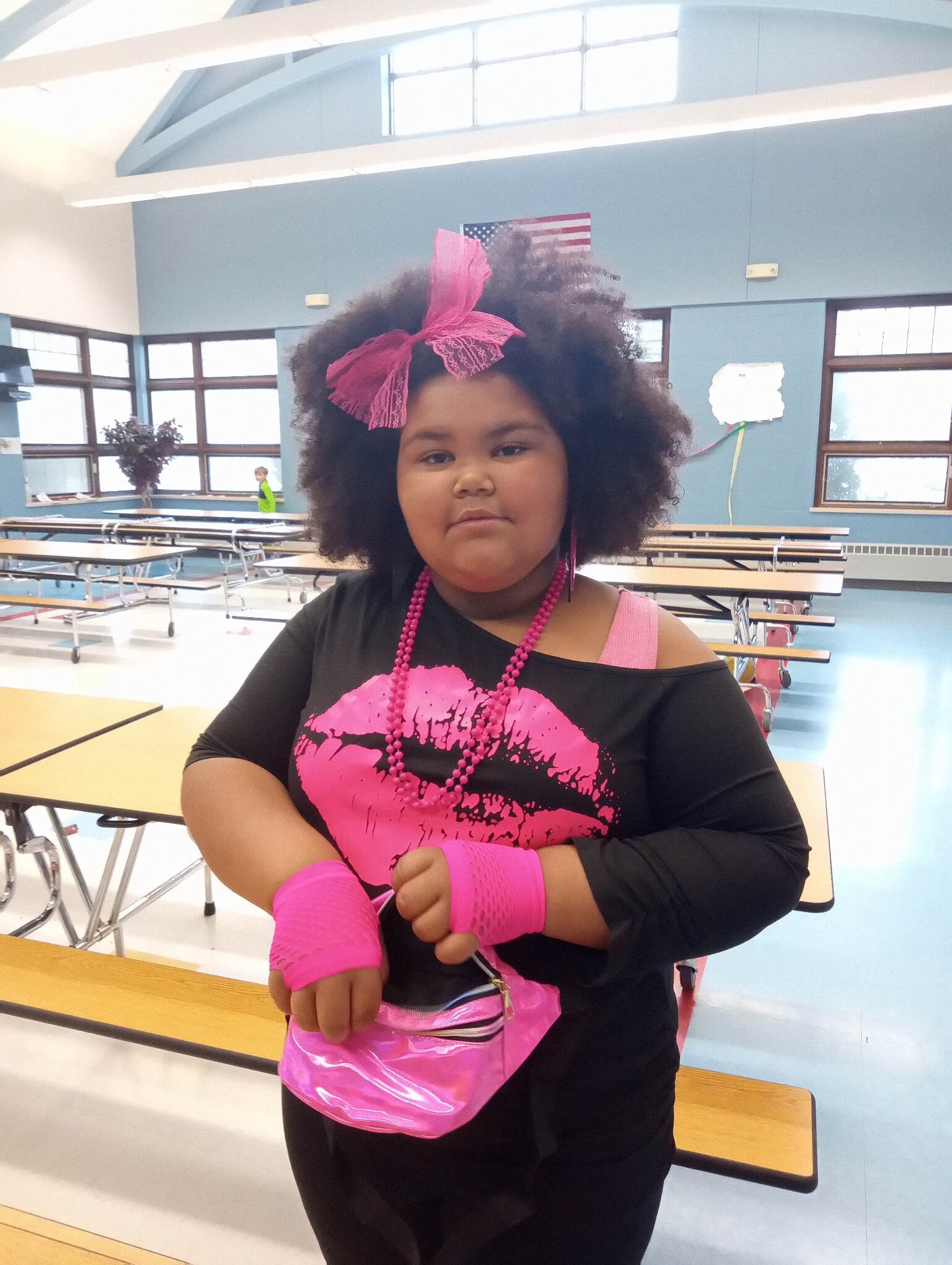 Girl in a school cafeteria wearing a large pink bow headband and pink accessories, posing near lunch tables.