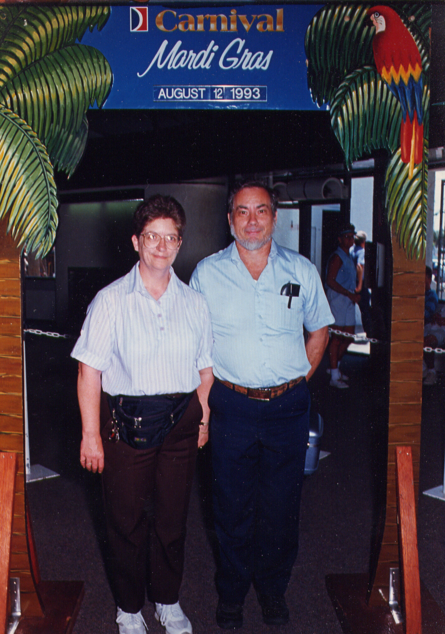 Two people pose together under a Mardi Gras Carnival sign showing the date August 12, 1993 in a tropical entrance setting with palm decorations.