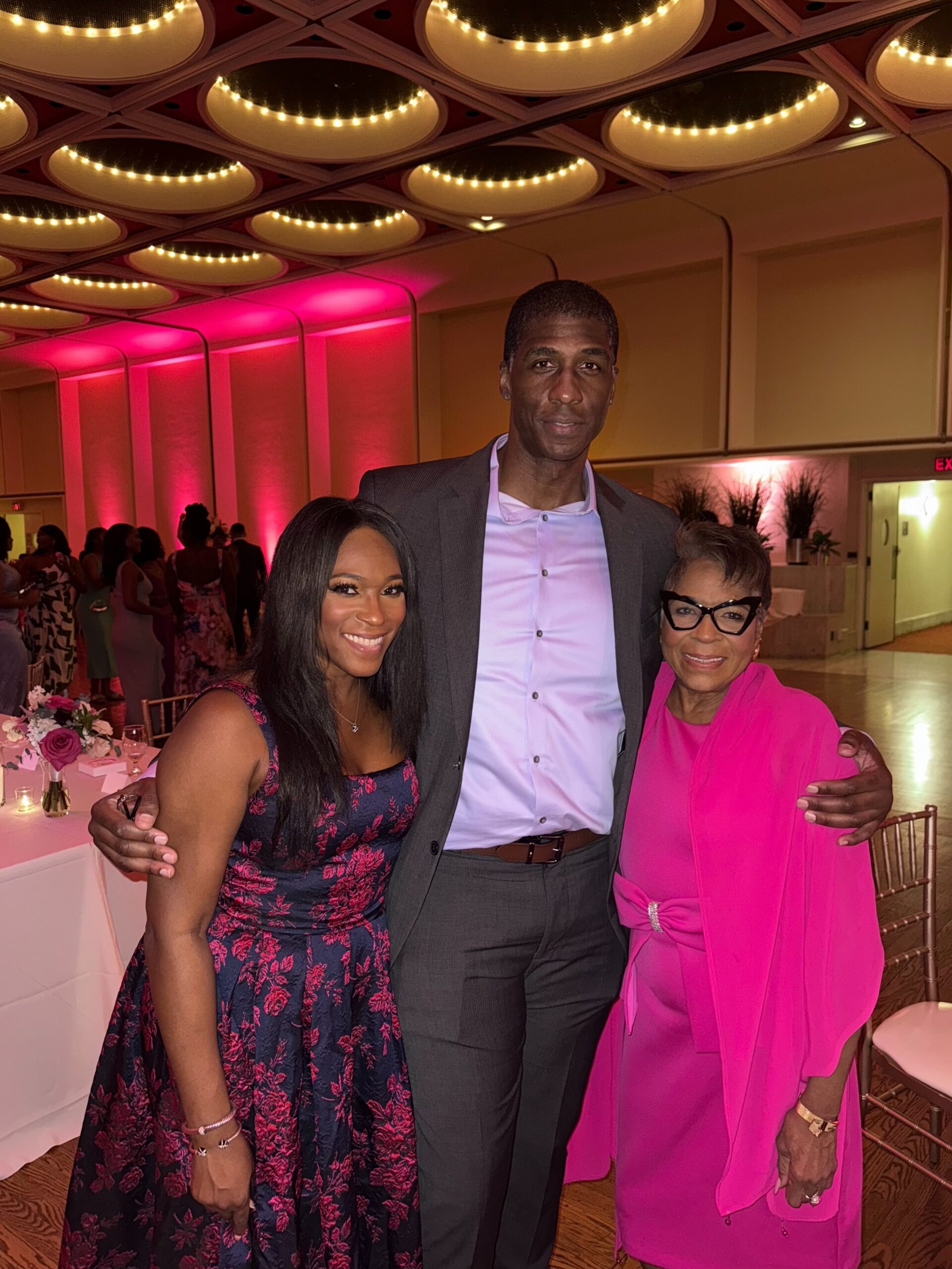 Three people posing together at a formal event, smiling under warm pink lighting inside a decorated venue.
