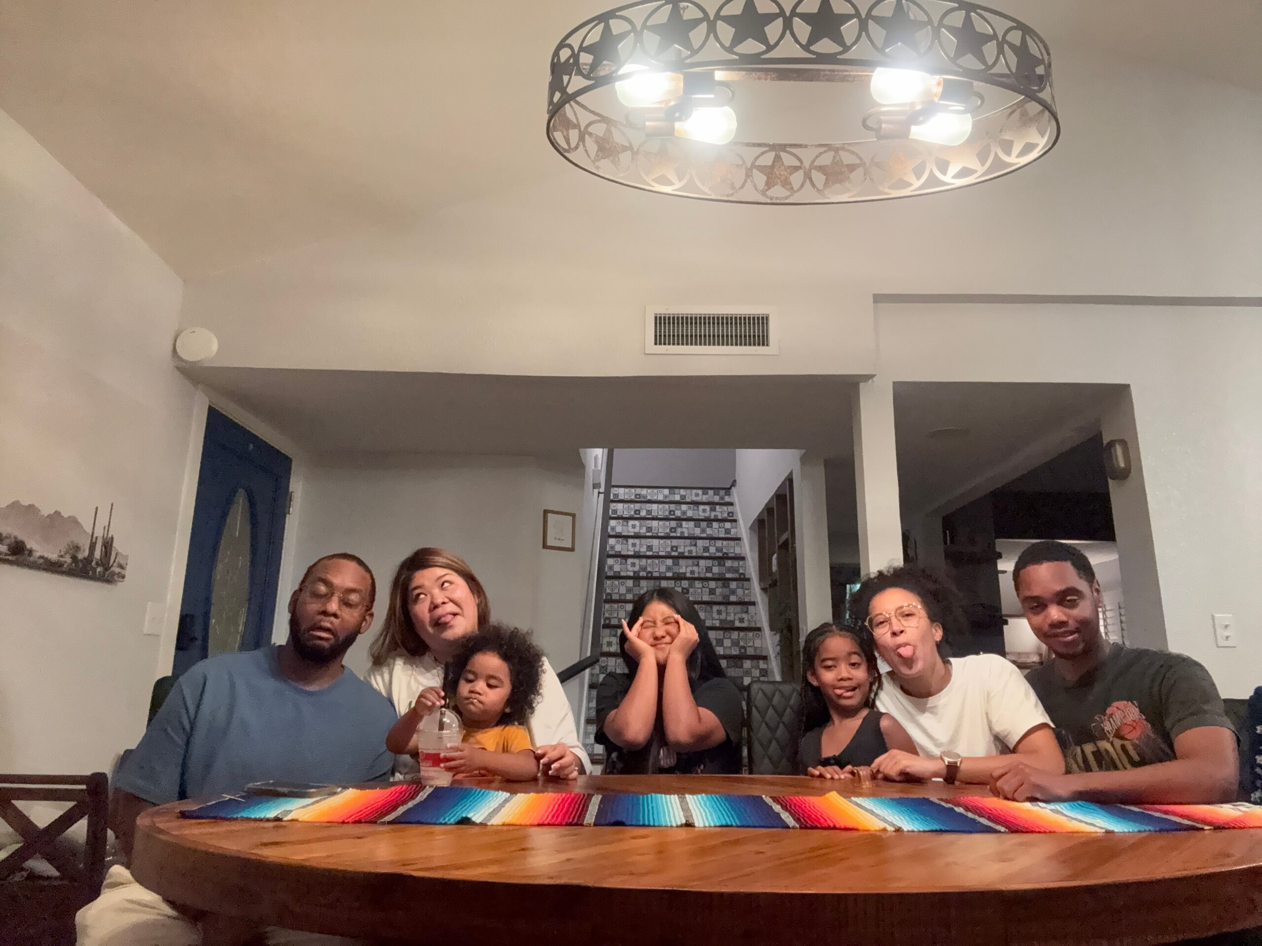 Group of people sitting around a wooden table indoors, smiling at the camera with a colorful striped table runner in view.”] ,