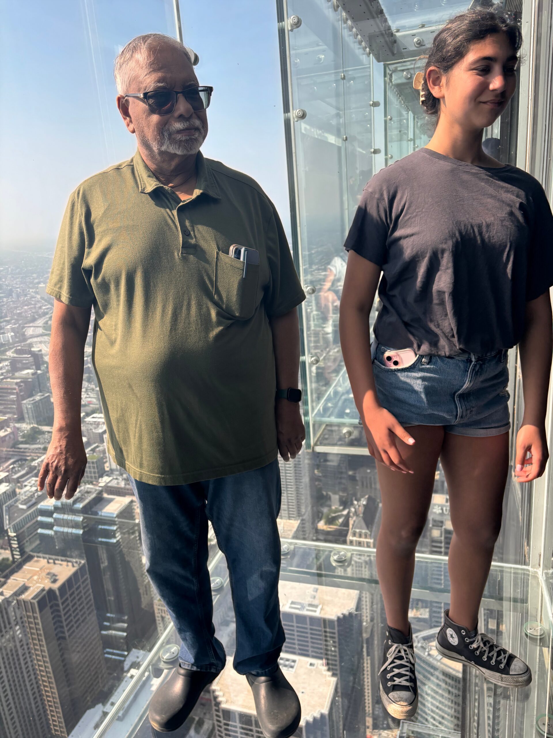 Older man in a green polo and sunglasses and a teenage girl in a black t-shirt stand on a glass observation deck high above a city.
