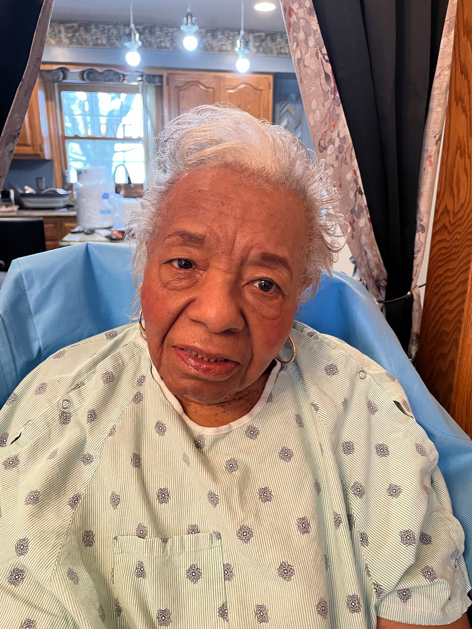 Close-up of an elderly woman in a light-striped hospital gown sitting in a blue chair, smiling at the camera. Background shows a kitchen area.