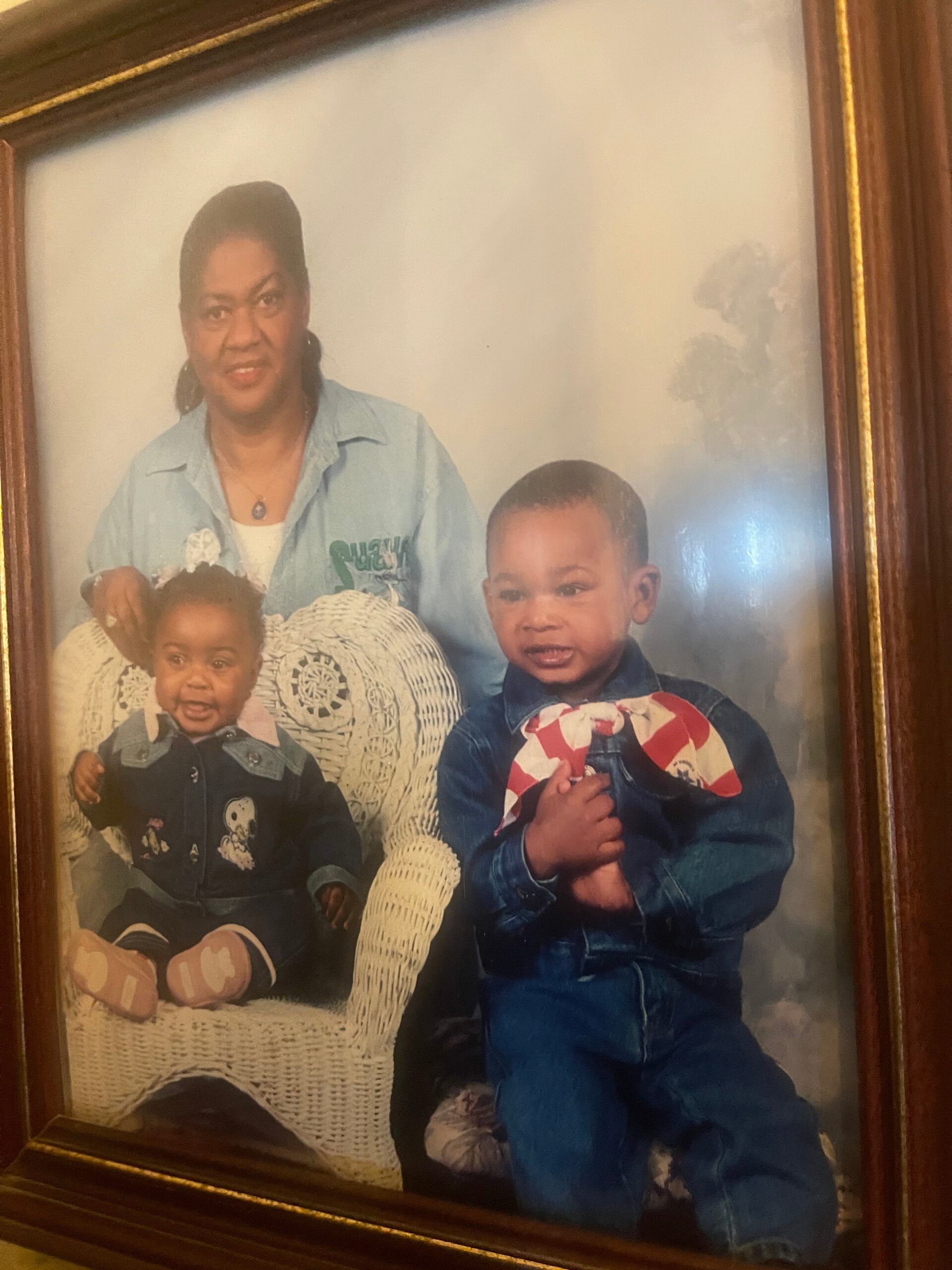 Mother standing behind two young children seated in a wicker chair, all posing for a family portrait.
