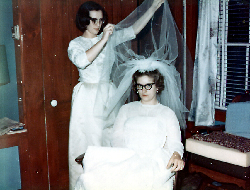 Two women in white wedding dresses; one sits in a chair wearing glasses while the other lifts a veil over her head.