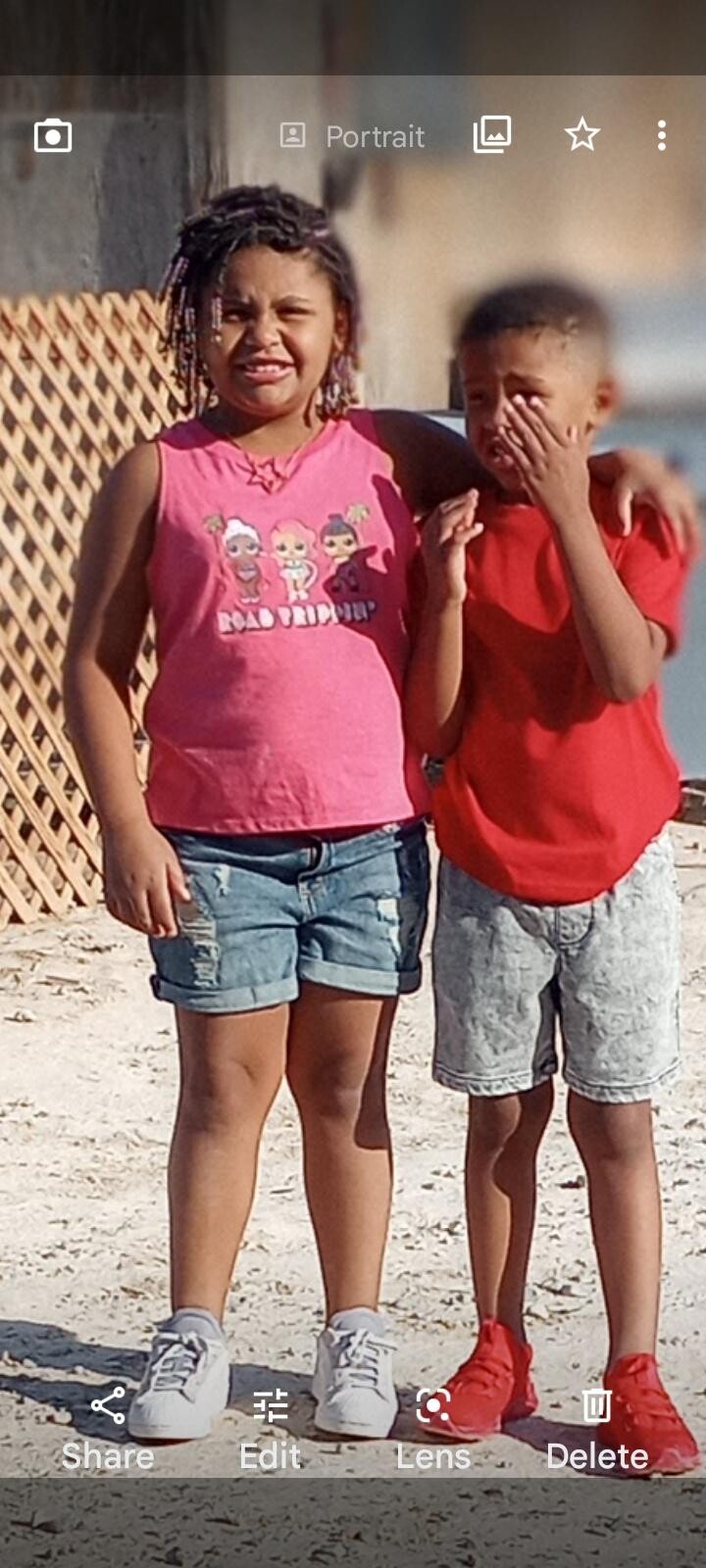 Two children stand together on a sandy beach, girl in a pink shirt and denim shorts, boy in red shirt and gray shorts, arms around each other, posing for a photo.