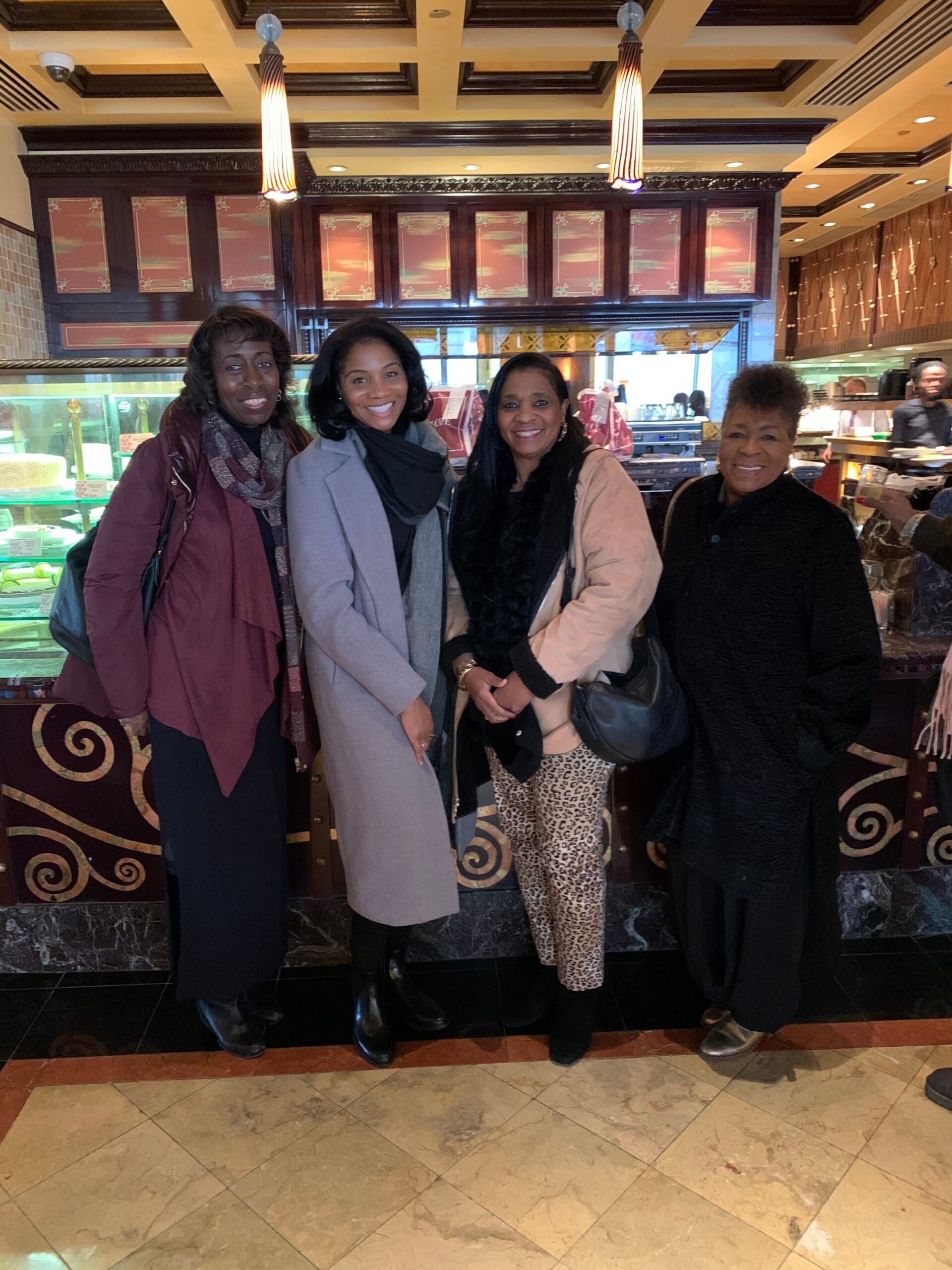 Four women smiling and posing together inside a bakery/c cafe with a glass pastry display behind them.