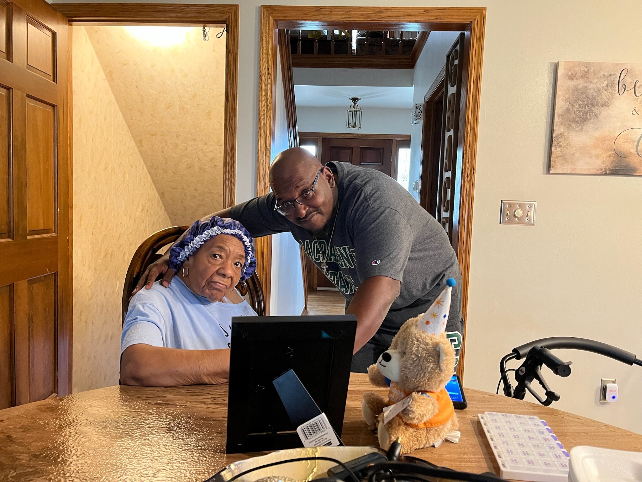 Elderly woman sitting at a table being assisted by a man, with a computer monitor and a stuffed bear nearby in a home dining area.