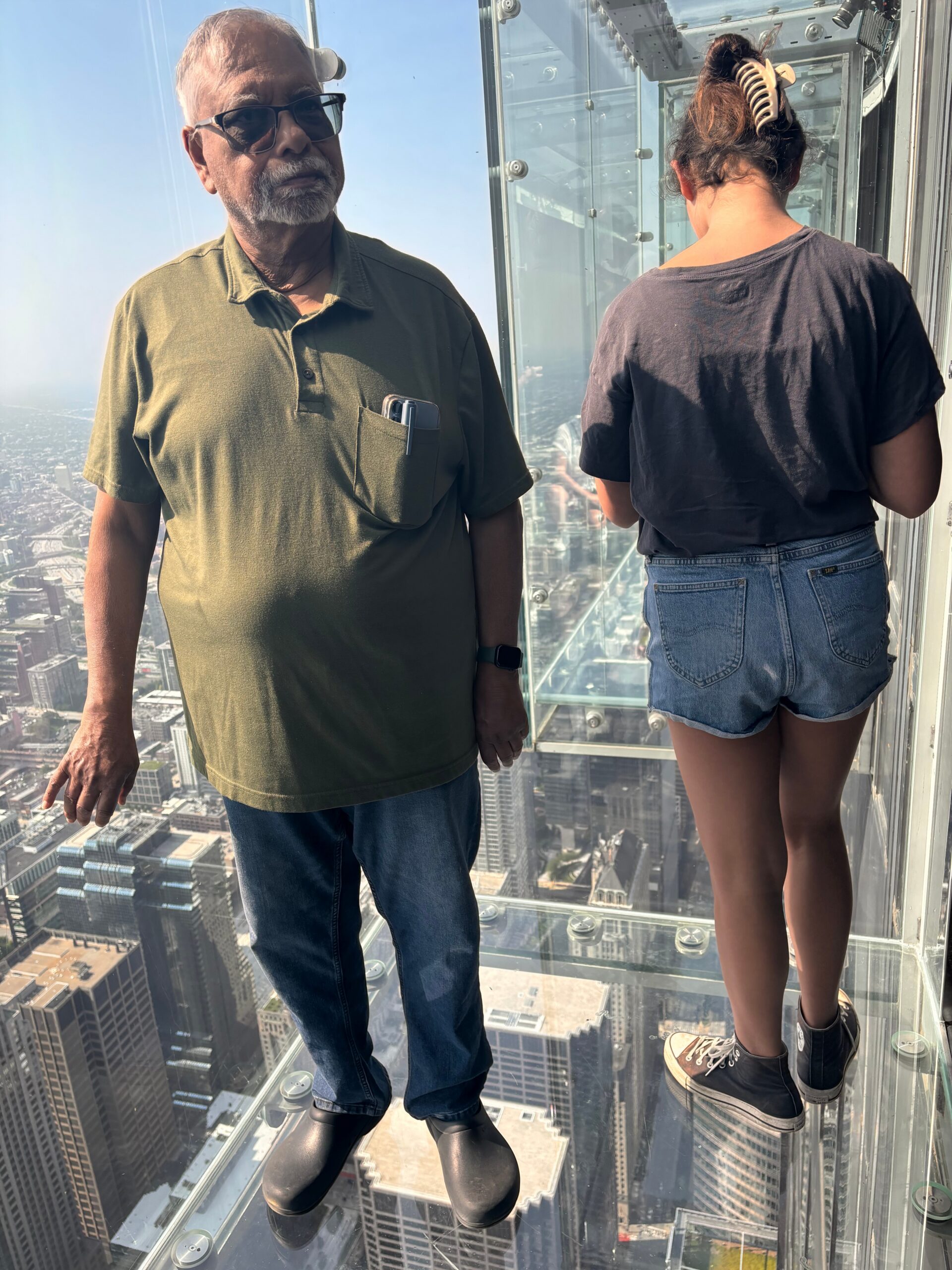 Two people stand on a glass observation deck high above a city skyline, looking down at skyscrapers below.