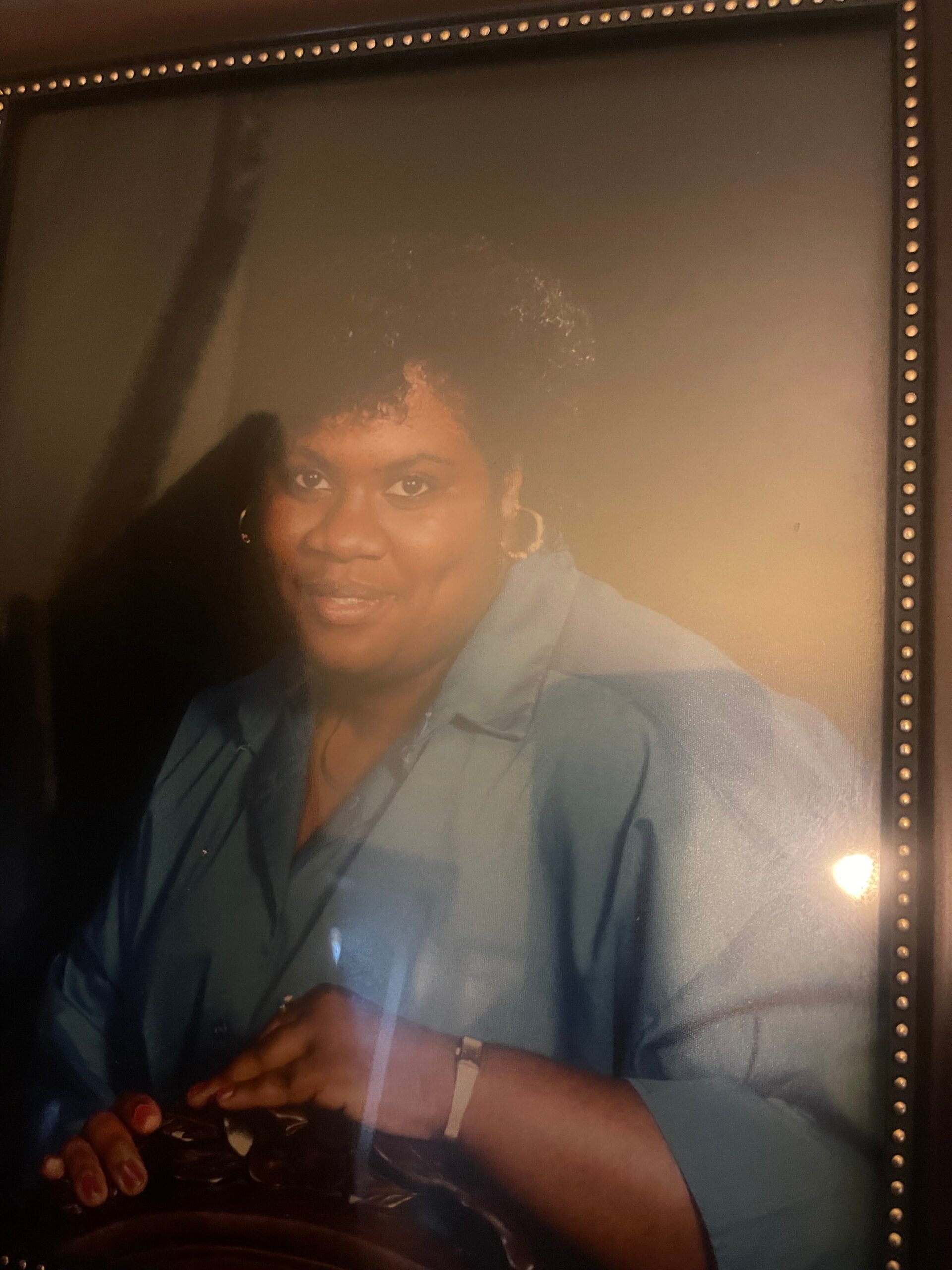 Portrait of a smiling Black woman wearing a light blue collared shirt, seated with hands folded in front of her.