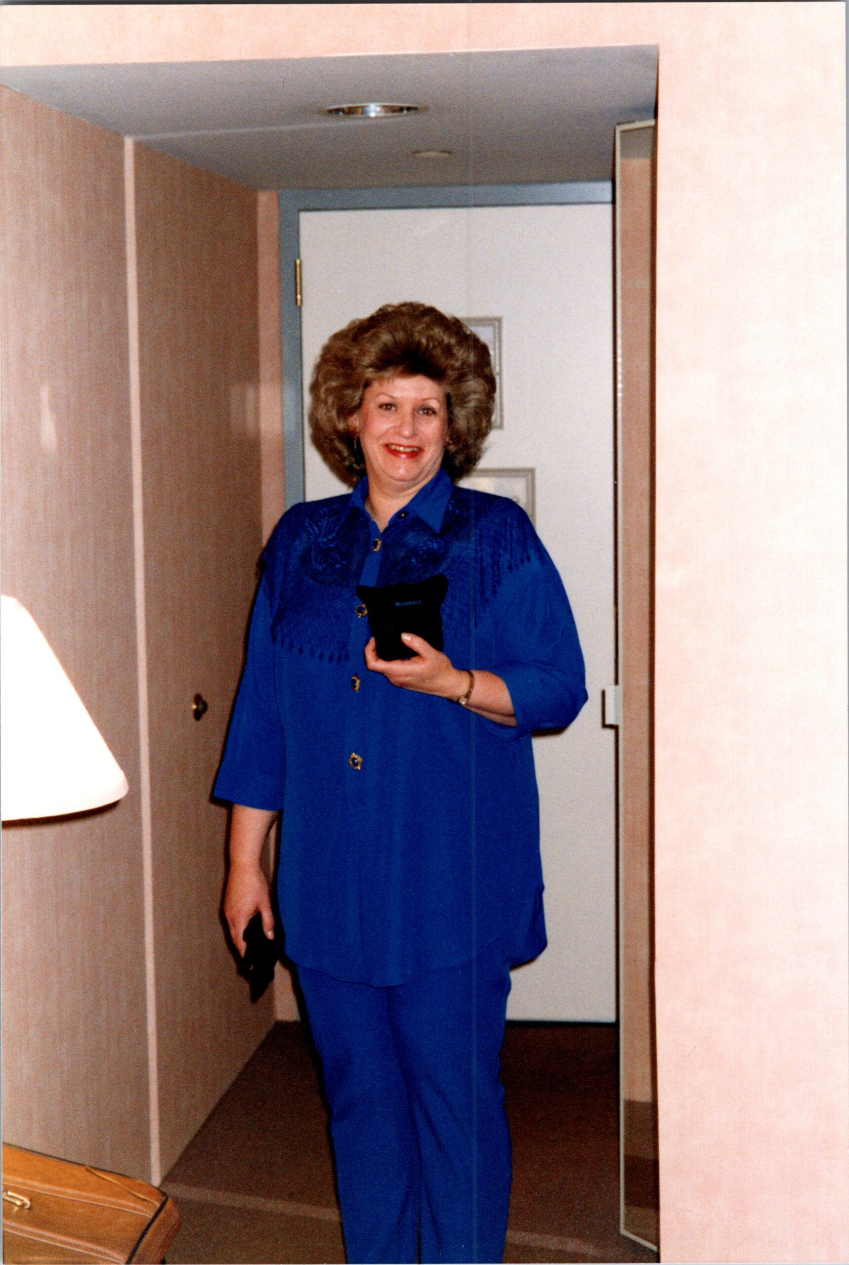 Woman in a royal blue blouse and pants taking a mirror selfie in a hotel room hallway with a door behind her.
