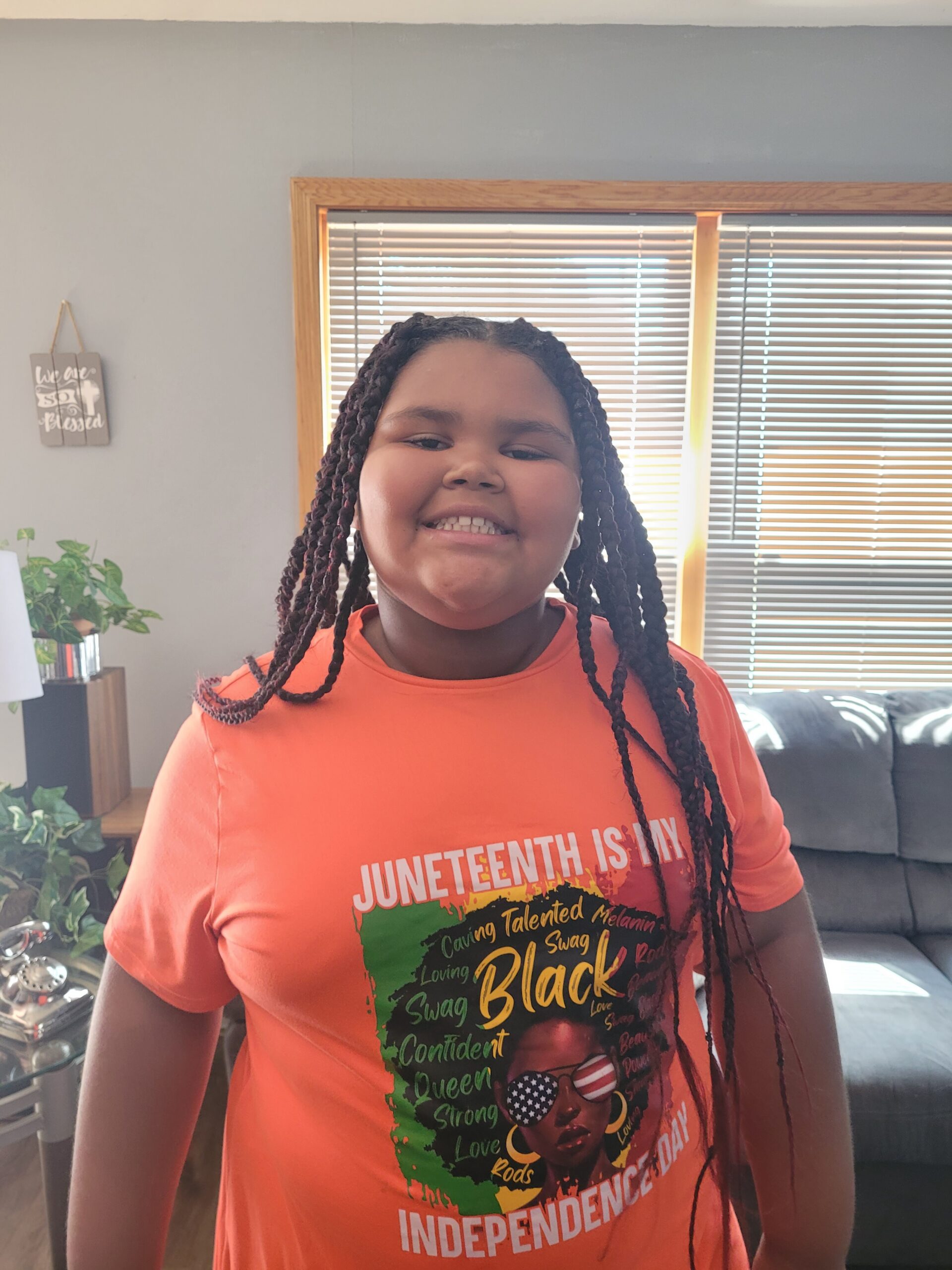 Girl with braided hair, wearing an orange Juneteenth shirt, smiling in a living room.
