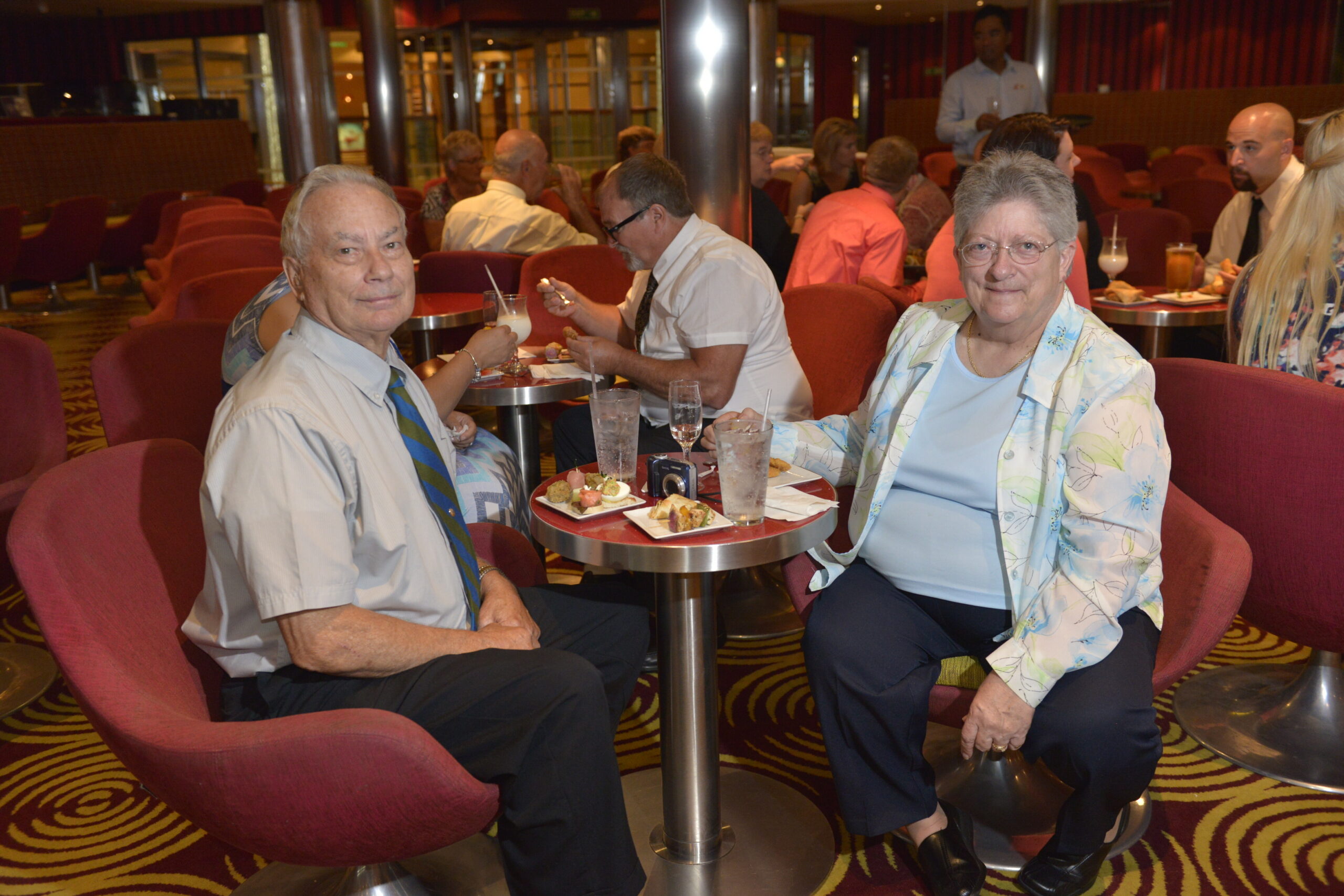 Two elderly people sit at a small round table in a red‑carpeted lounge, enjoying a meal and drinks together while others chat in the background.