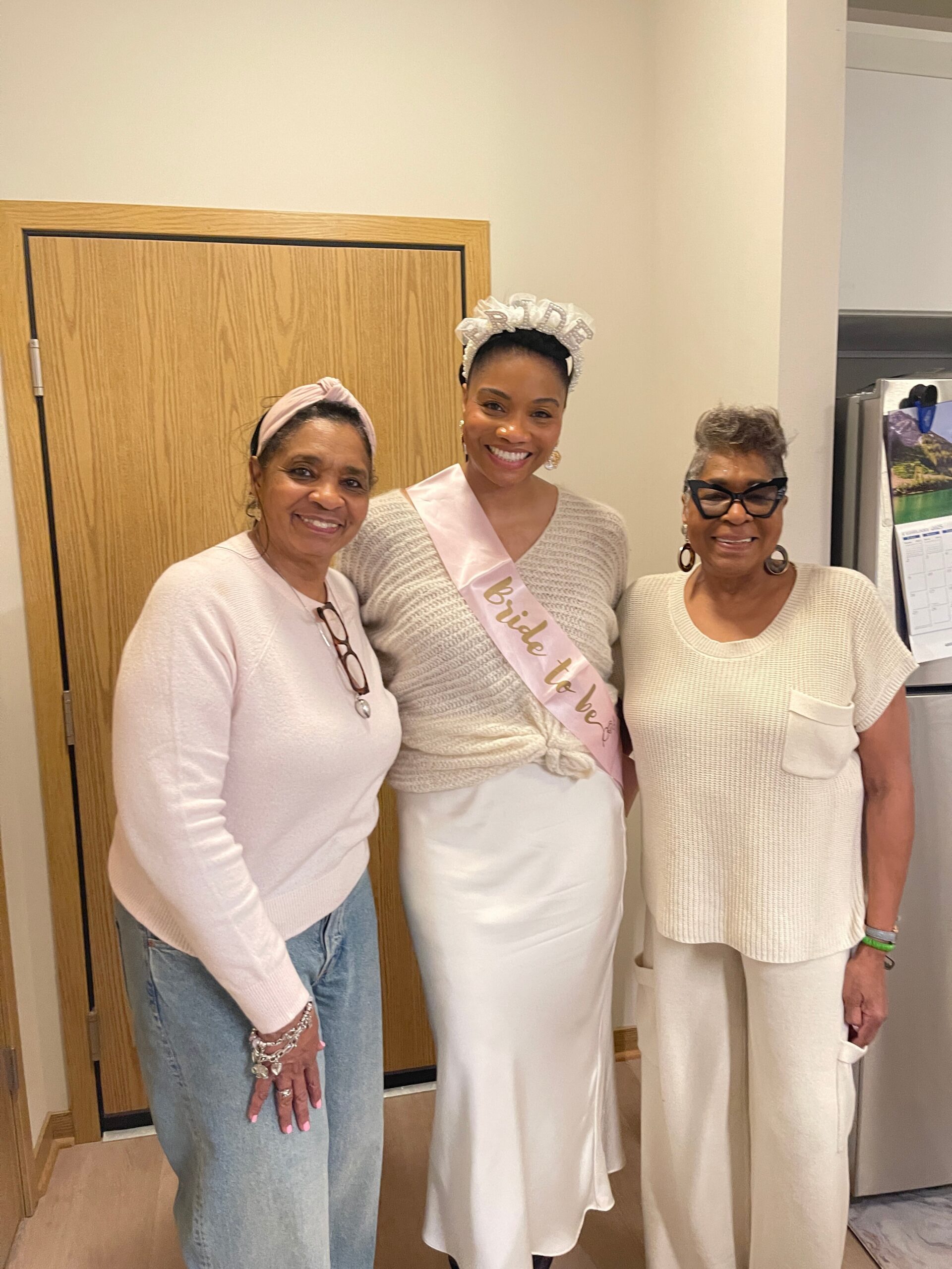 Three women posing together; center bride-to-be in white with a 'Bride to be' sash and crown, flanked by her friends.
