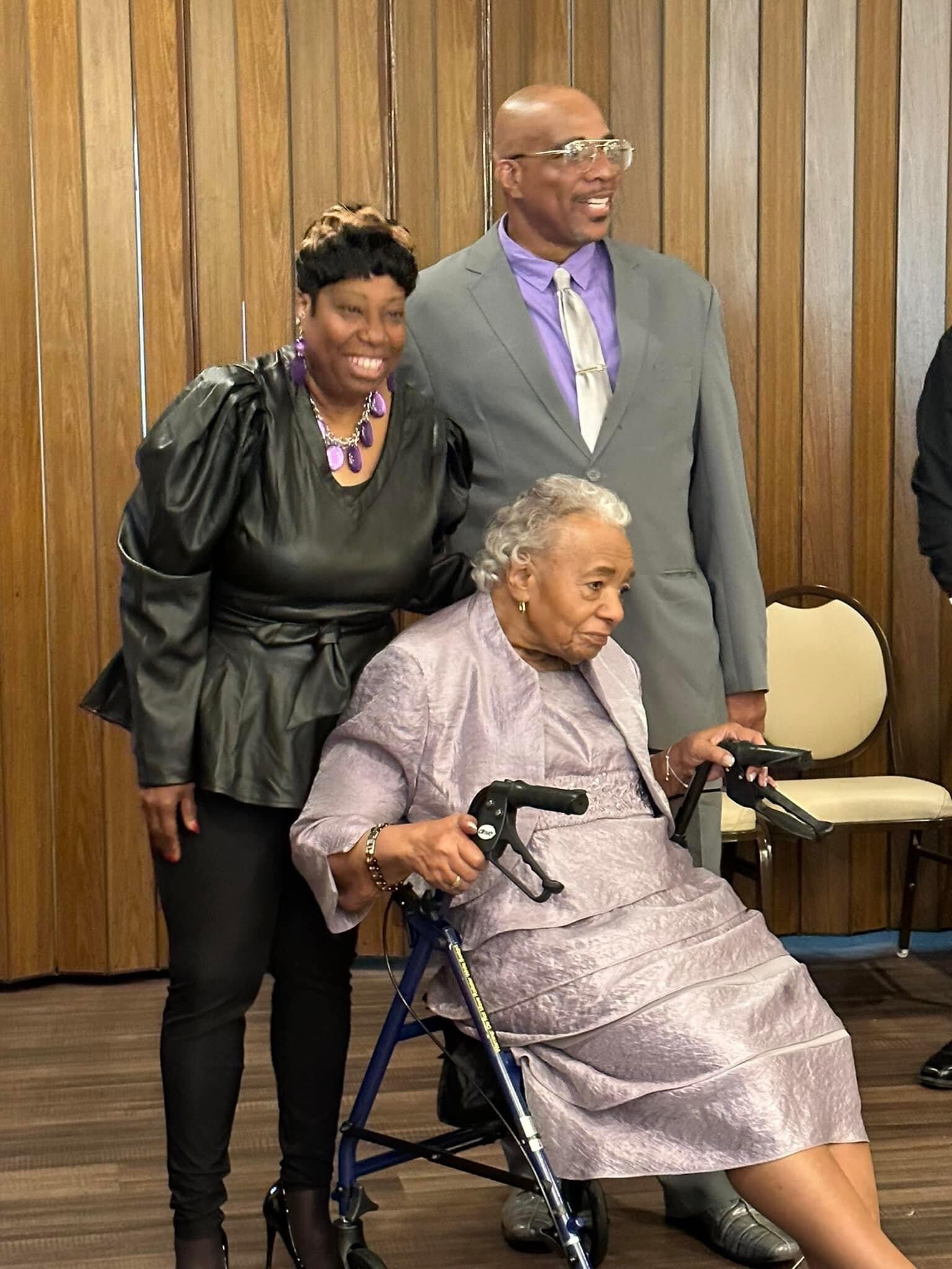 Smiling elderly woman in a lavender dress sits in a walker, with a man in a gray suit and lavender shirt standing behind her and a woman in black beside them against wooden paneling.