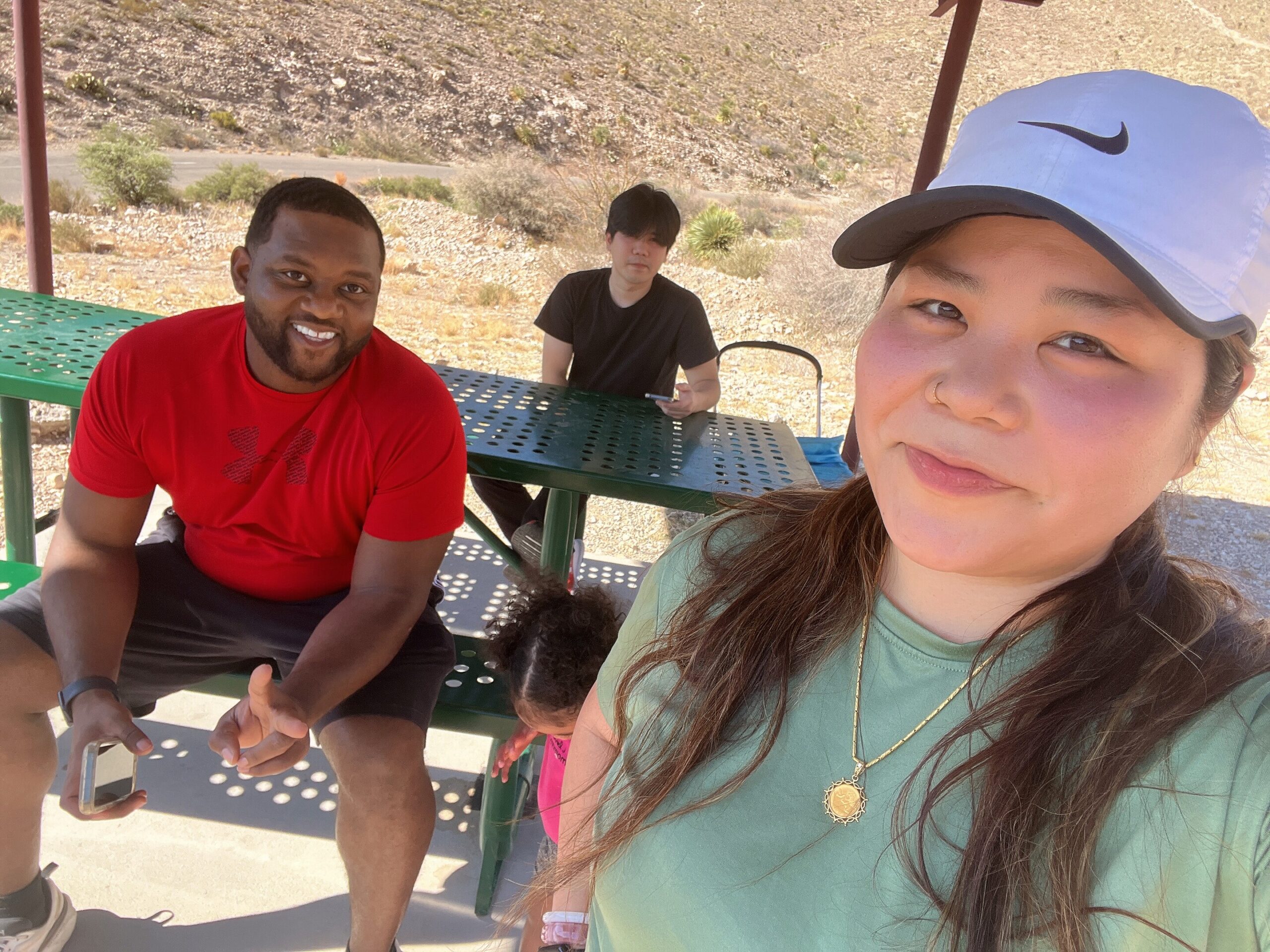 Three friends sit at a green perforated picnic table outdoors, smiling at the camera with a dry, hilly background.