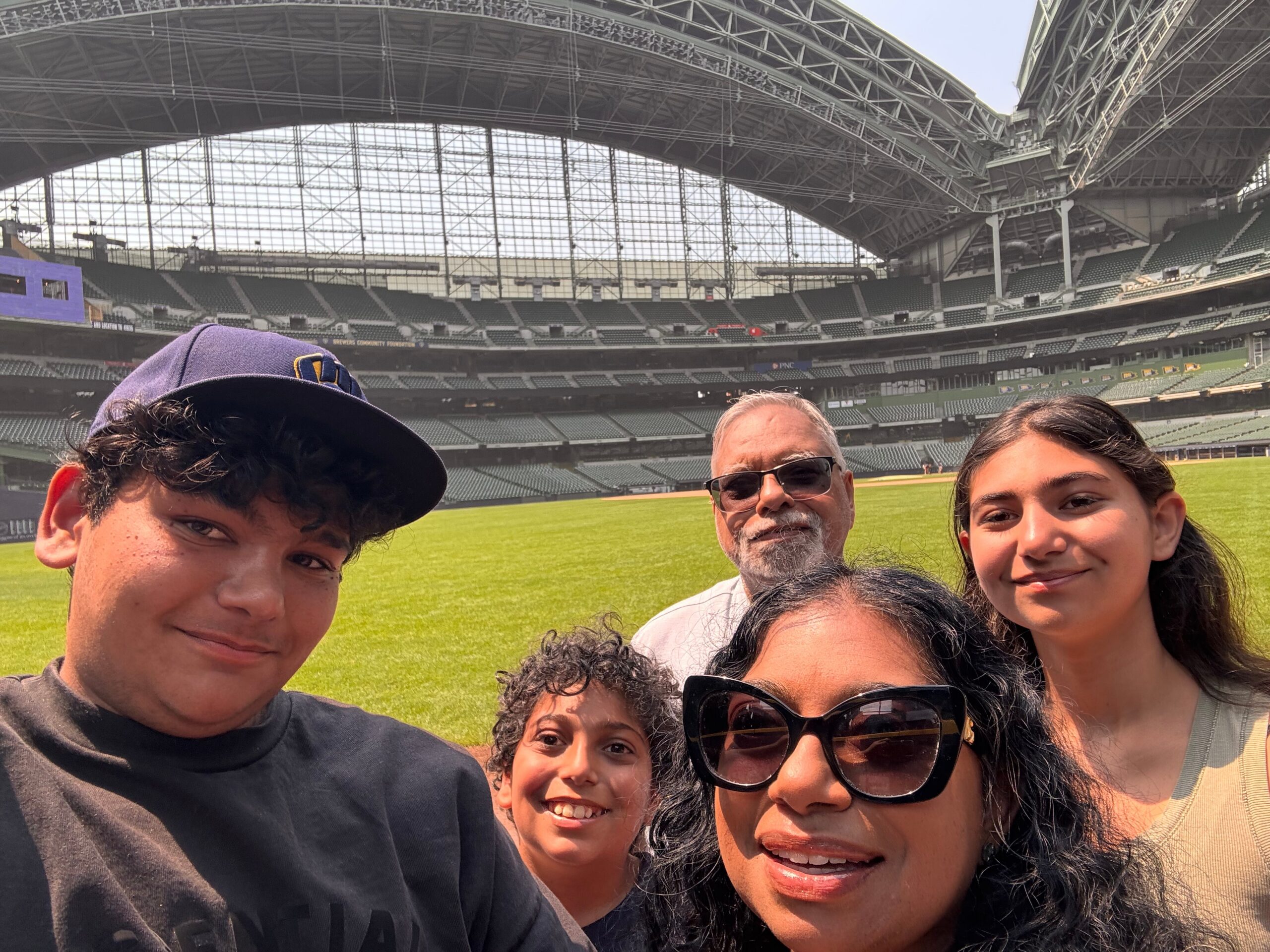 Group of friends taking a selfie on a baseball field inside a large empty stadium with a steel roof structure behind them.