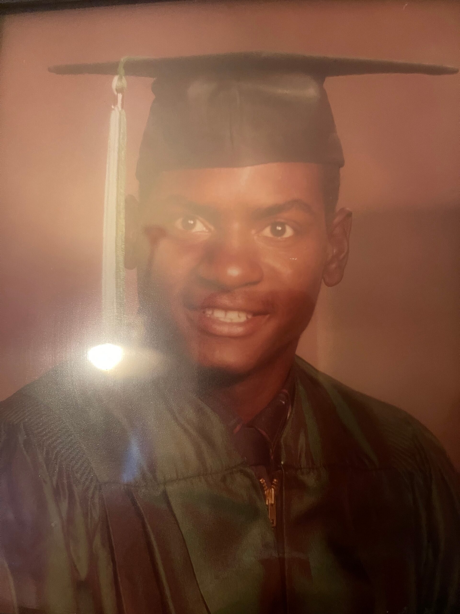 Portrait of a young man wearing a graduation cap (mortarboard) and a jacket, smiling at the camera against a pink backdrop.