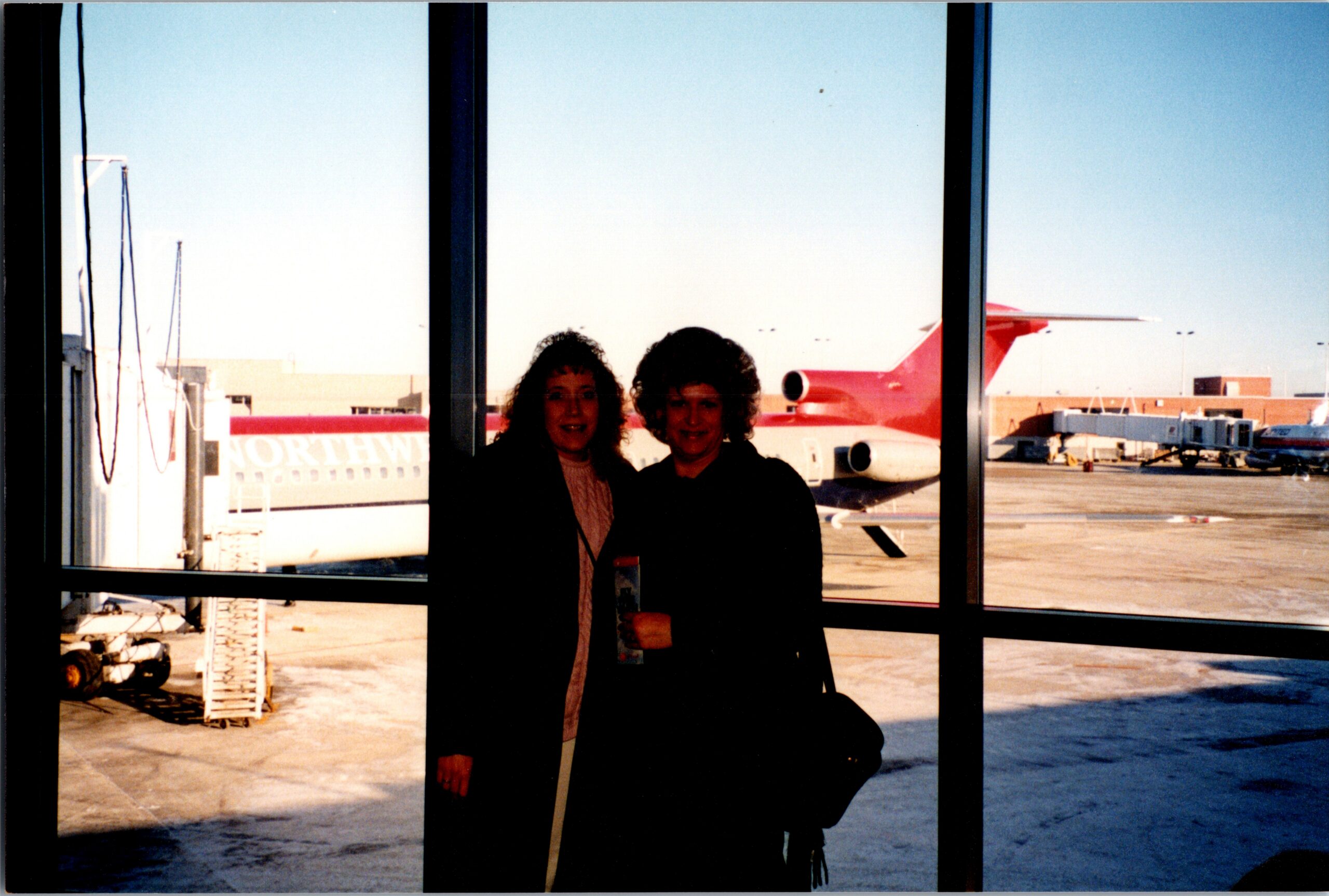 Two people posing inside an airport terminal with a red airplane visible on the tarmac outside the windows.