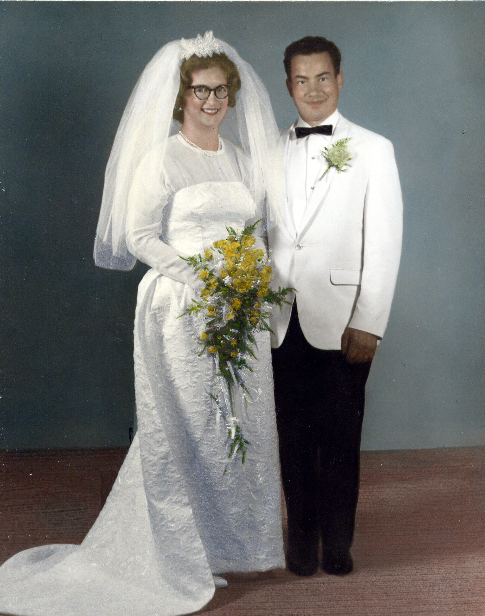 Bride in a white lace gown and veil holds a yellow bouquet, standing beside a groom in a white suit with a black bow tie.