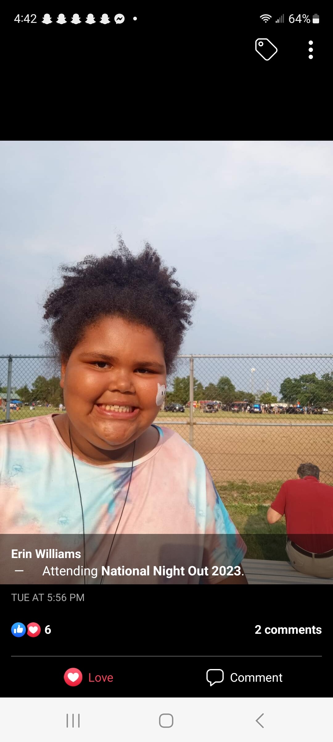 Smiling girl with curly hair in a pastel tie-dye shirt at an outdoor event behind a chain-link fence.