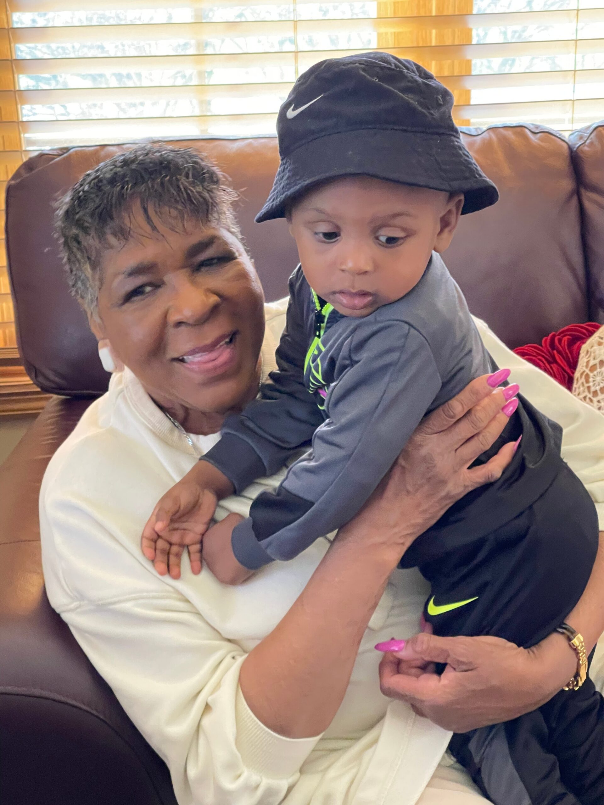 Grandmother smiling while holding a toddler on a brown couch with window blinds in the background.