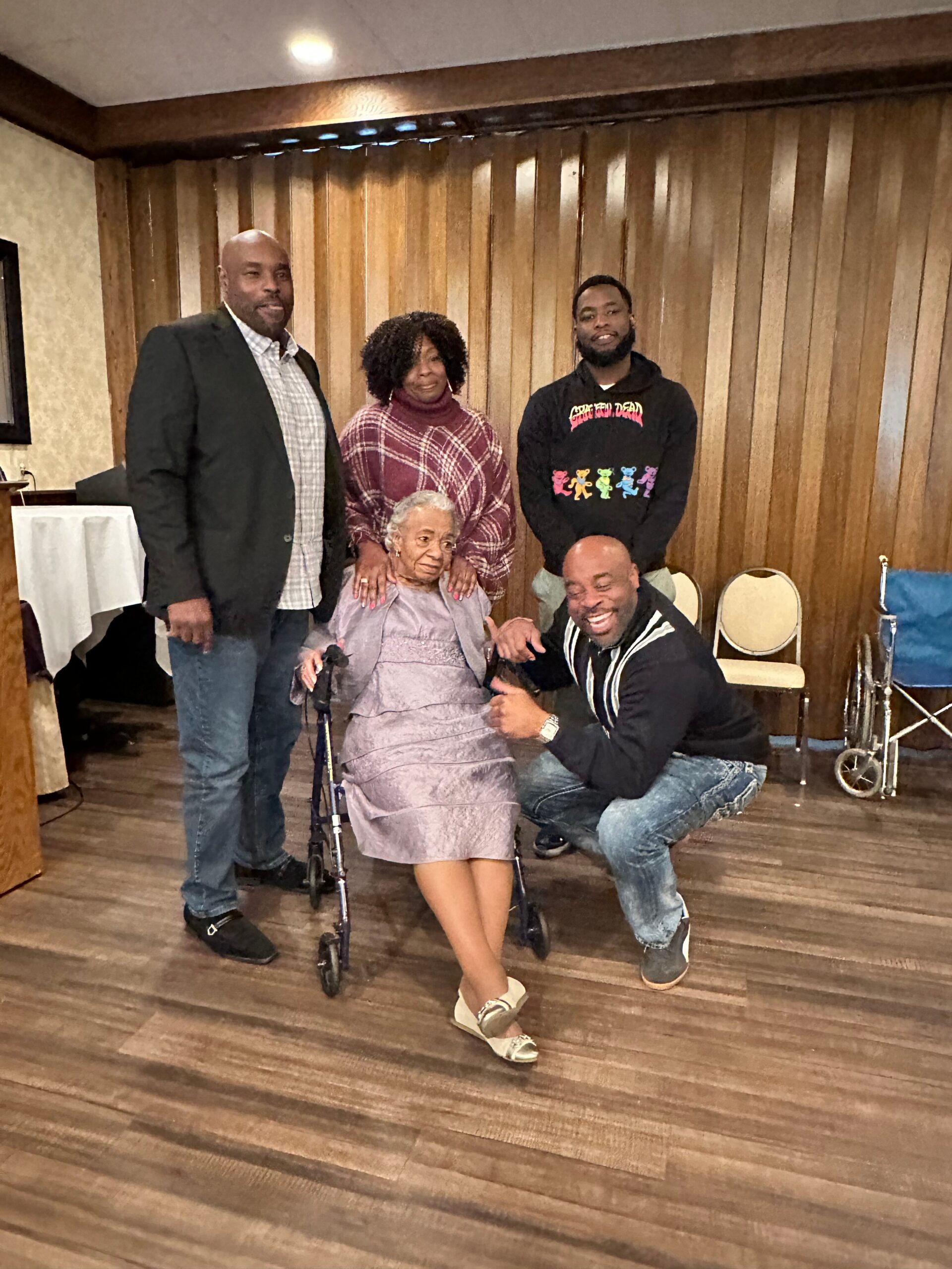 Group of five people posing indoors; an elderly woman in a wheelchair is center, surrounded by four smiling relatives standing or kneeling nearby.
