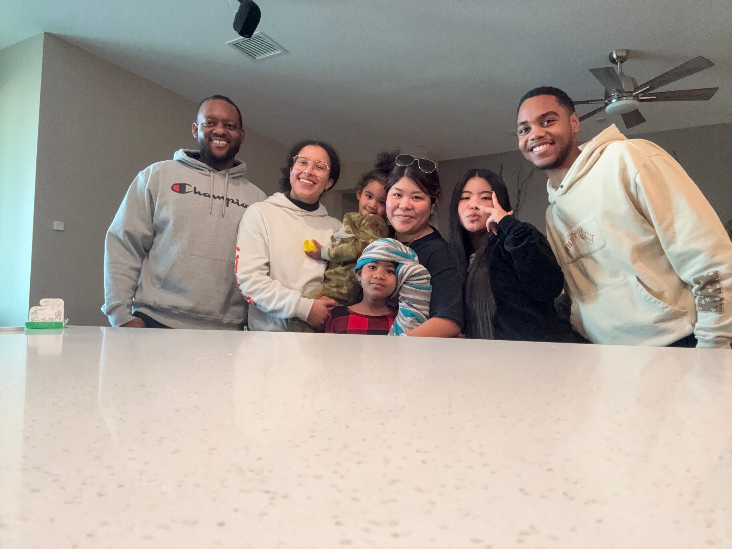 Seven friends and kids pose for a casual group selfie indoors, leaning on a white counter and smiling at the camera.
