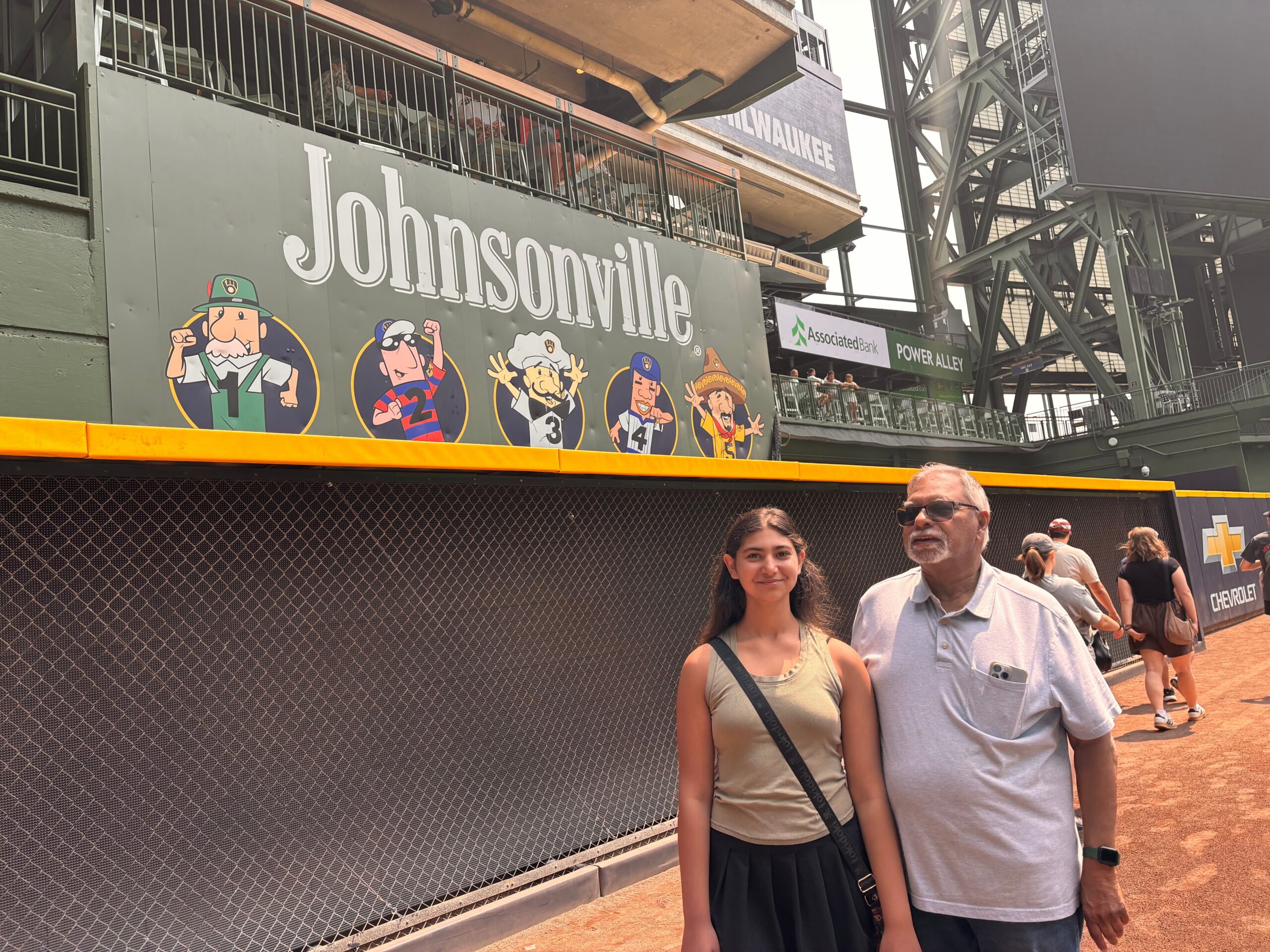 Two people pose at a stadium concourse beside a large Johnsonville sign featuring cartoon chefs.