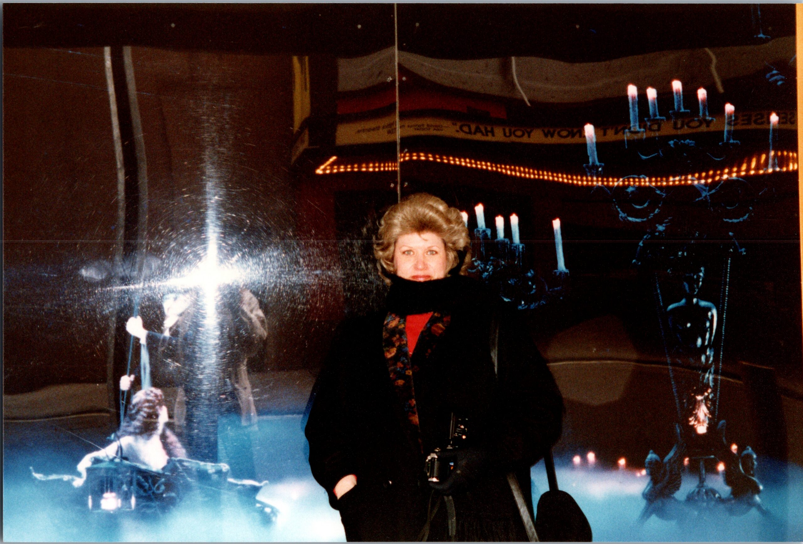 Smiling woman in a dark coat and scarf standing in a dim room with candle-like lights and reflections around her.