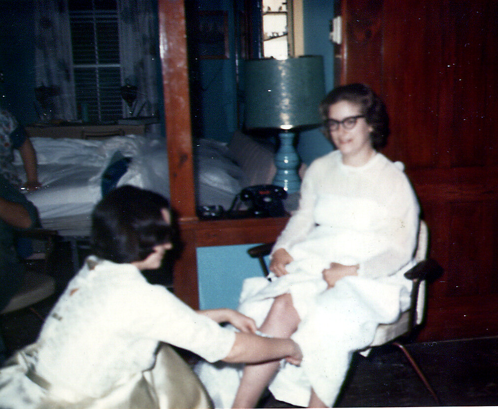 Woman in a white dress sits in a chair as another person in white robes tends to her bare foot nearby.