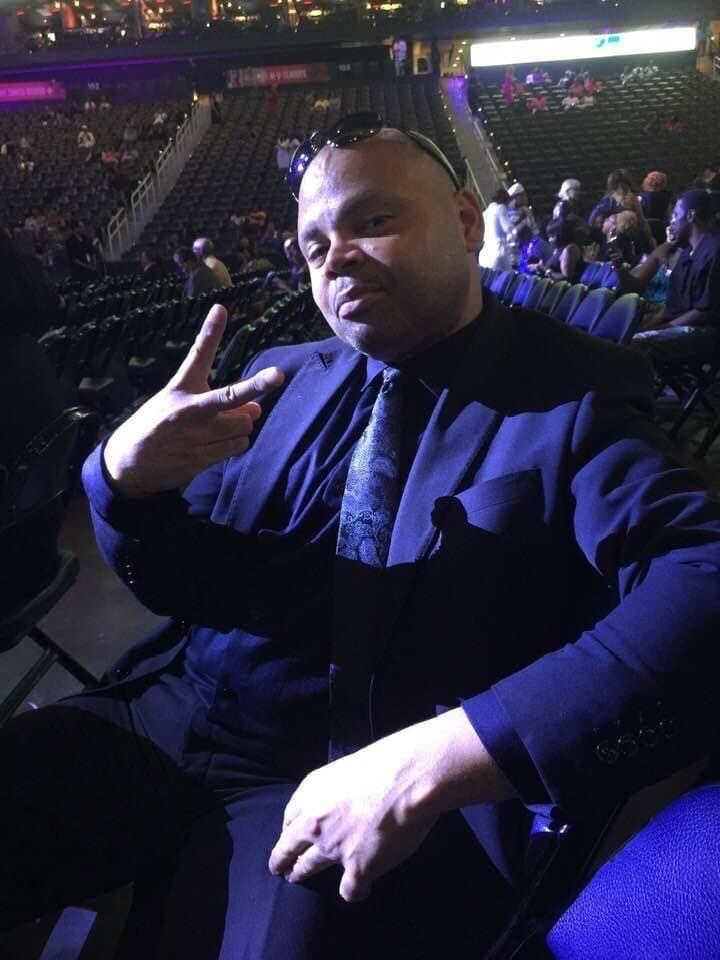 Man in a blue suit and tie sits in an arena, flashing a peace sign at the camera.