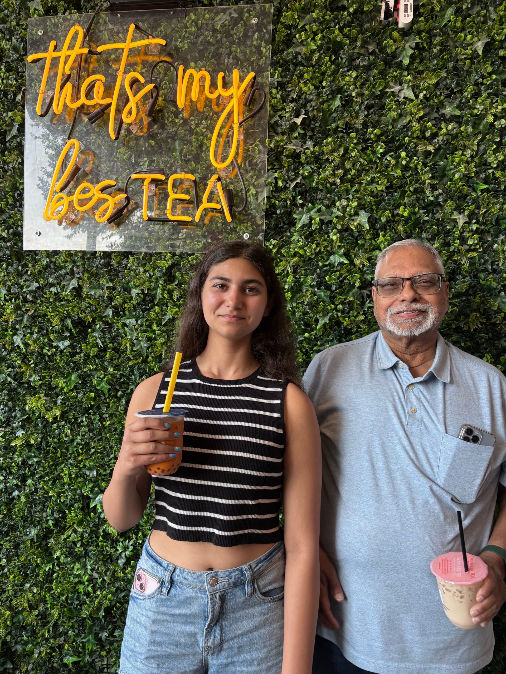 Young woman and older man pose in front of a green ivy wall with a neon sign that says, 'That's my best tea'; she holds a bubble tea drink.