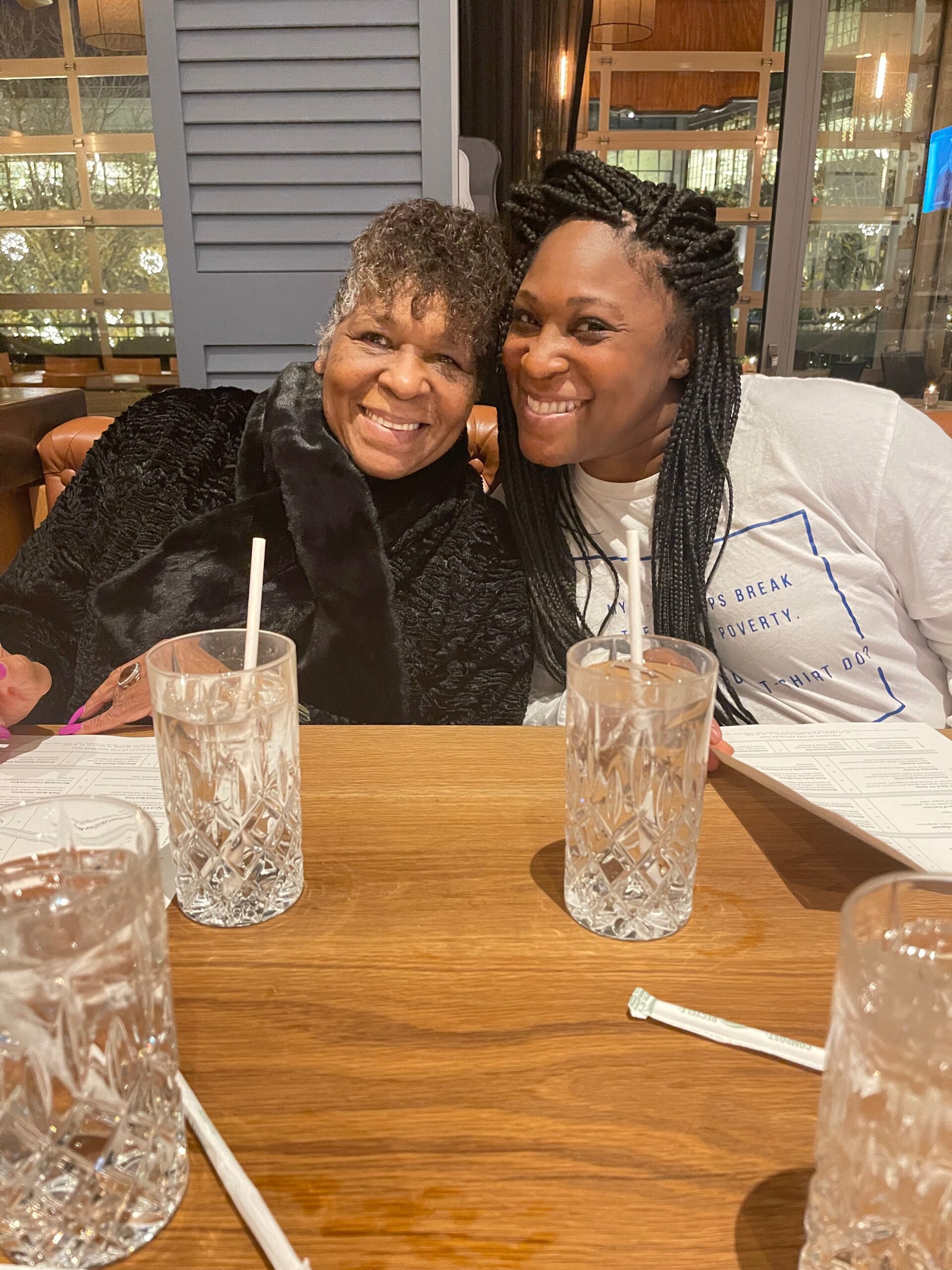 Two women sit closely at a restaurant table, smiling at the camera with drinks in crystal glasses and straws in the foreground, a menu visible on the table.