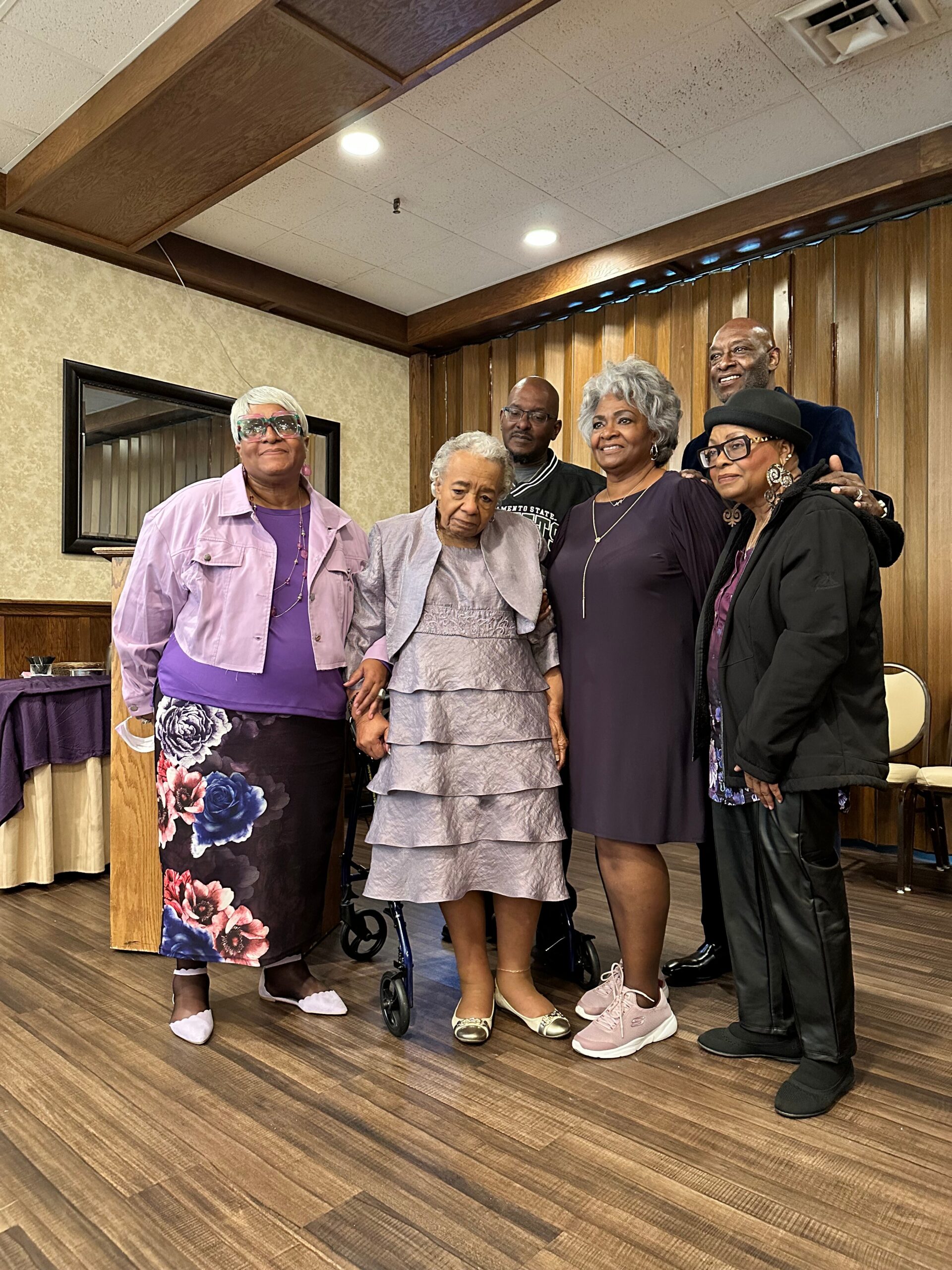 A group of six smiling adults posing together indoors, with one in a walker, in front of wood-paneled wall.