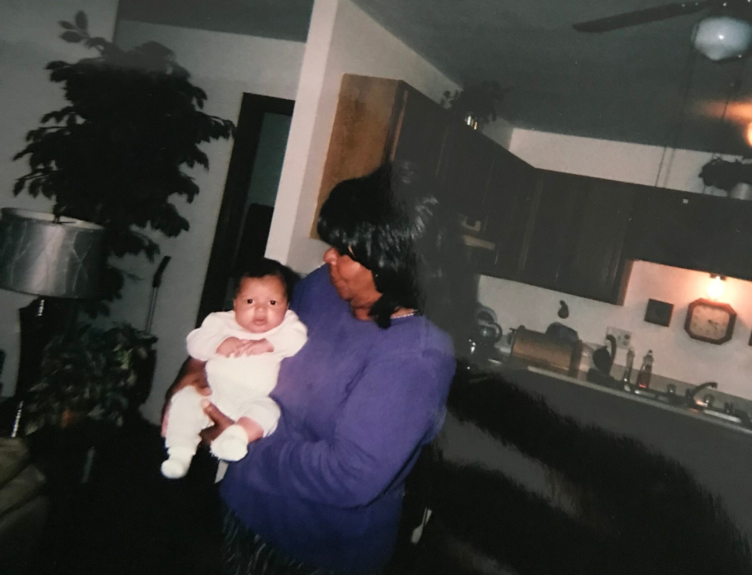 A person wearing a purple top holds a baby in white clothing indoors, with a large plant and kitchen area in the background.
