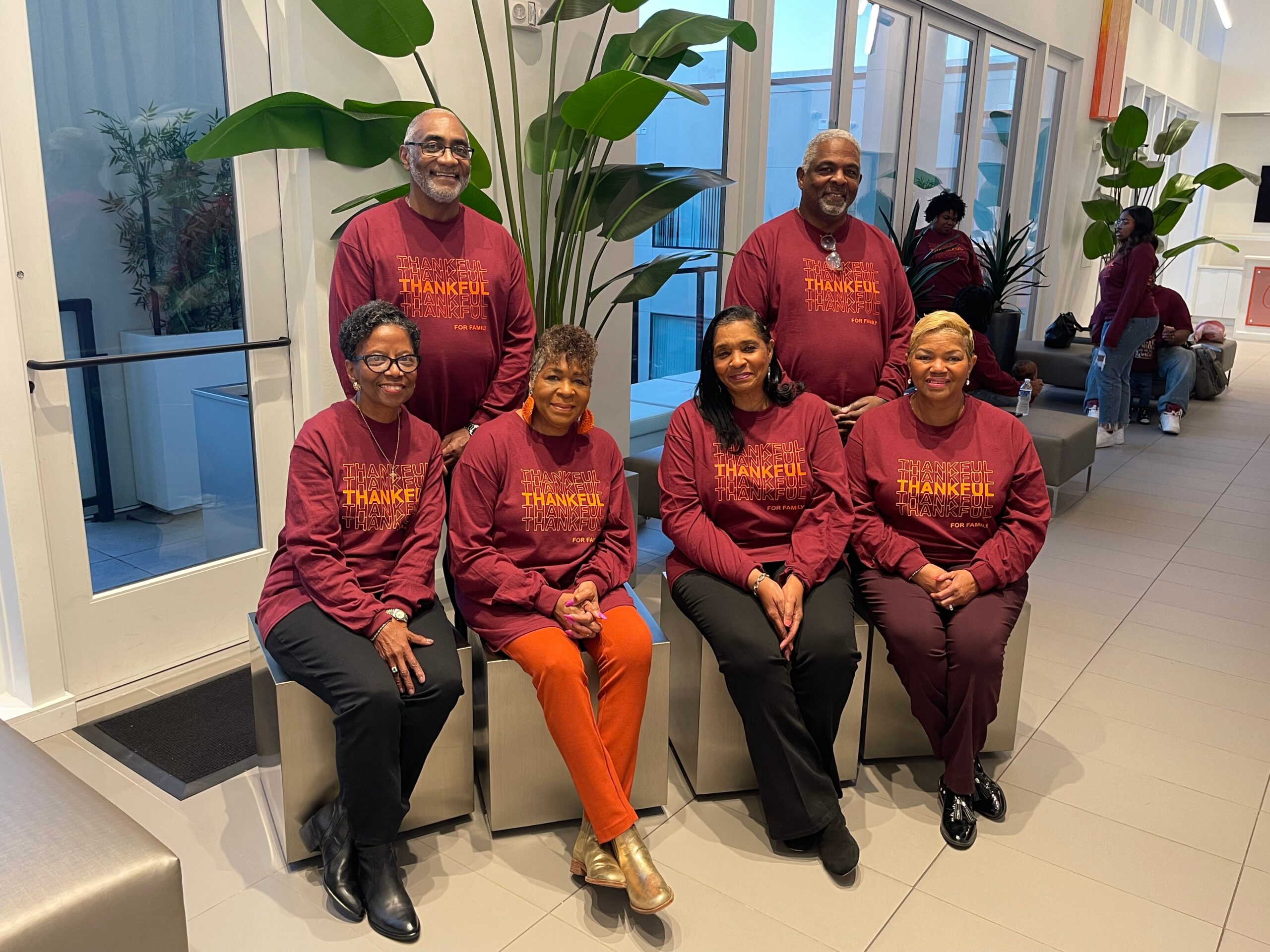 Six adults in maroon shirts posing together in a modern lobby with large plants in the background.