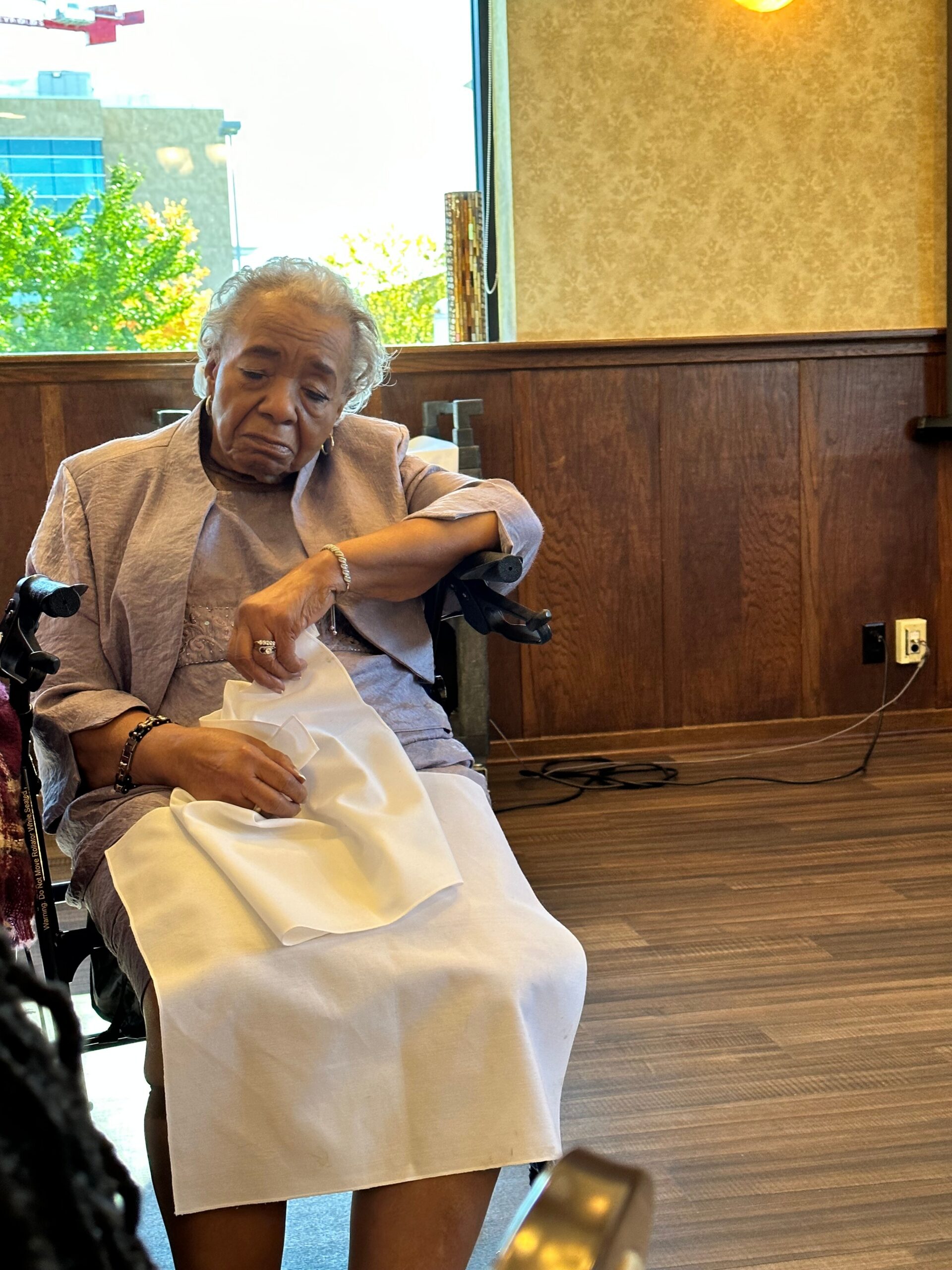 Older person in a wheelchair resting with a white napkin on their lap, indoors beside a wooden wall paneling.