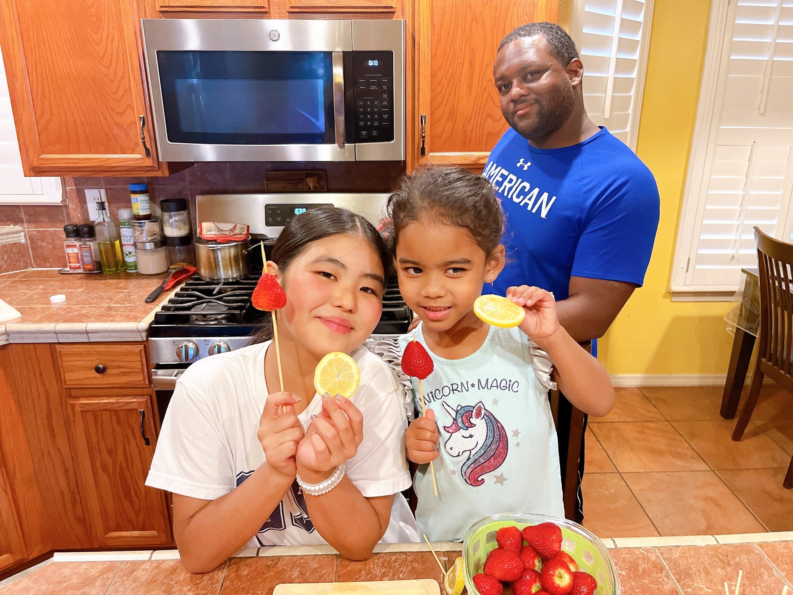 Two smiling girls hold fruit skewers (orange slices and strawberries) in a kitchen, with a man in a blue shirt smiling in the background.