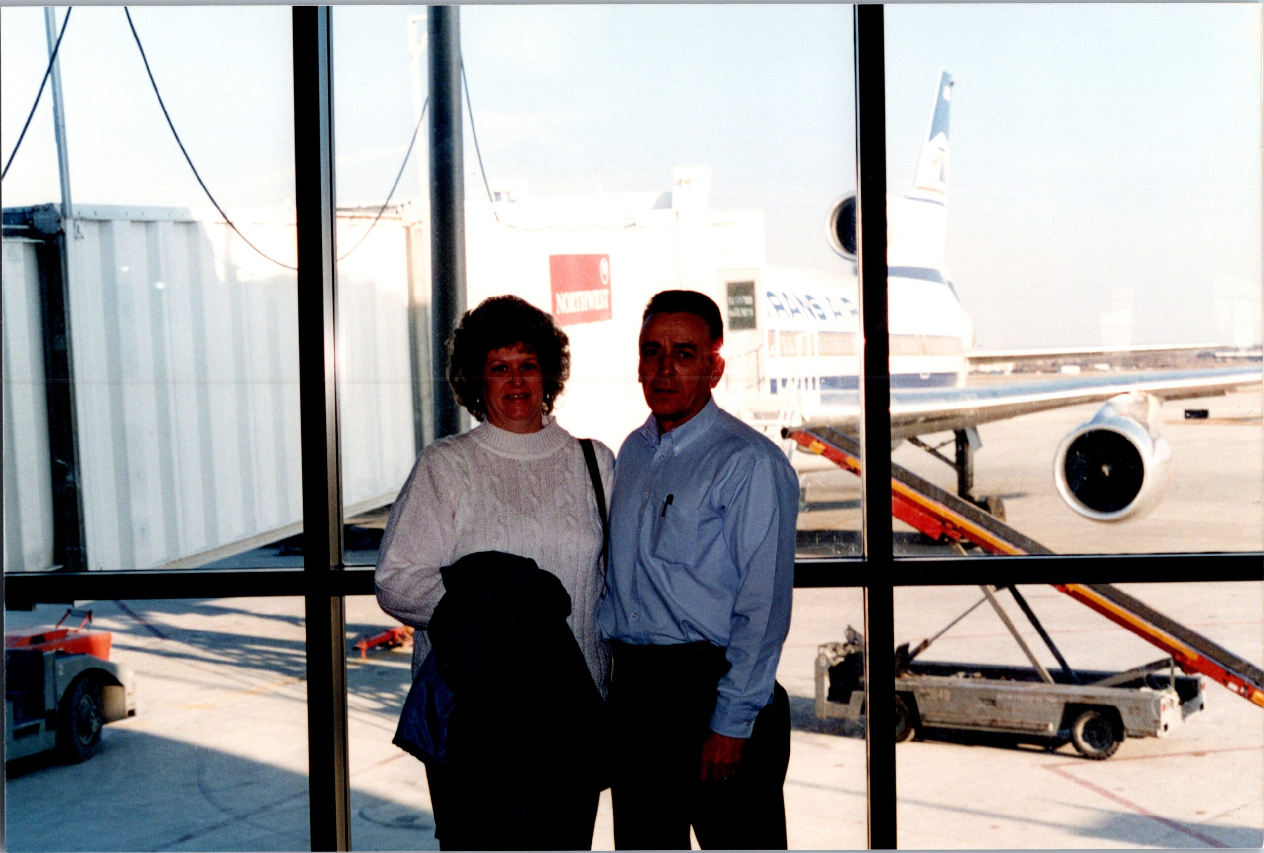 Two people stand by a large window at an airport gate, with a plane and jet bridge outside behind them.