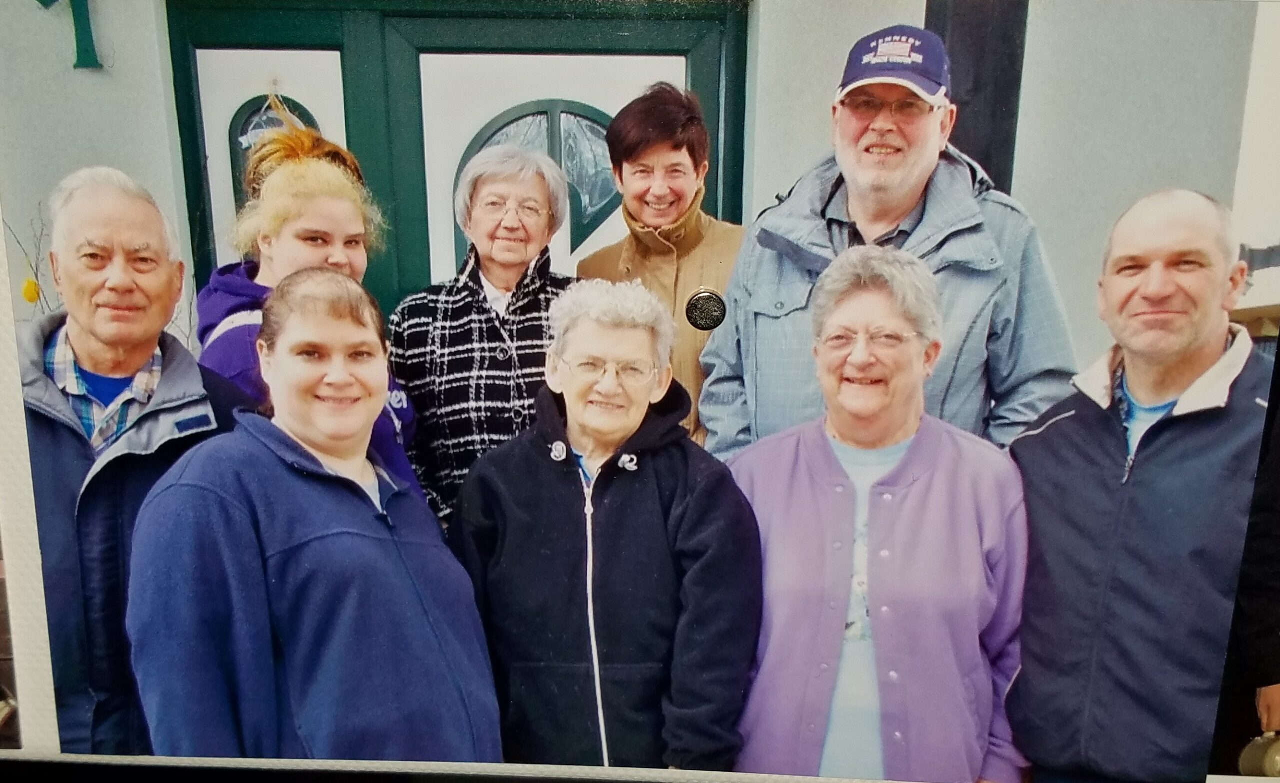 Group of nine adults posing closely together outside, smiling, in front of a green double door.