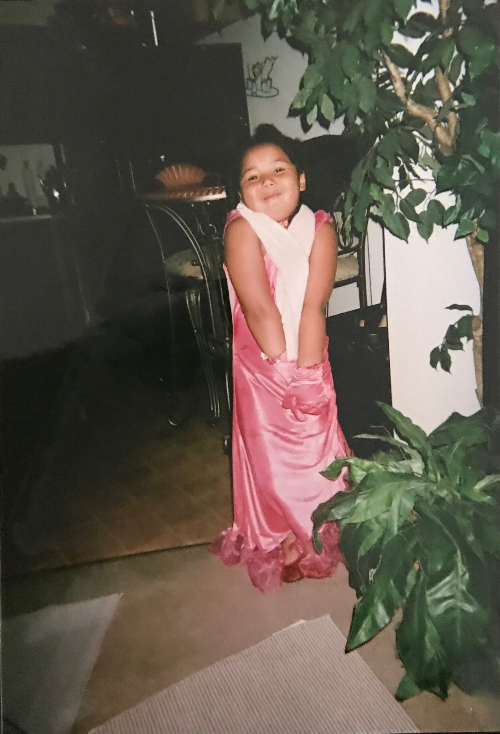 Young girl in a pink dress posing with a large leafy plant, smiling indoors.