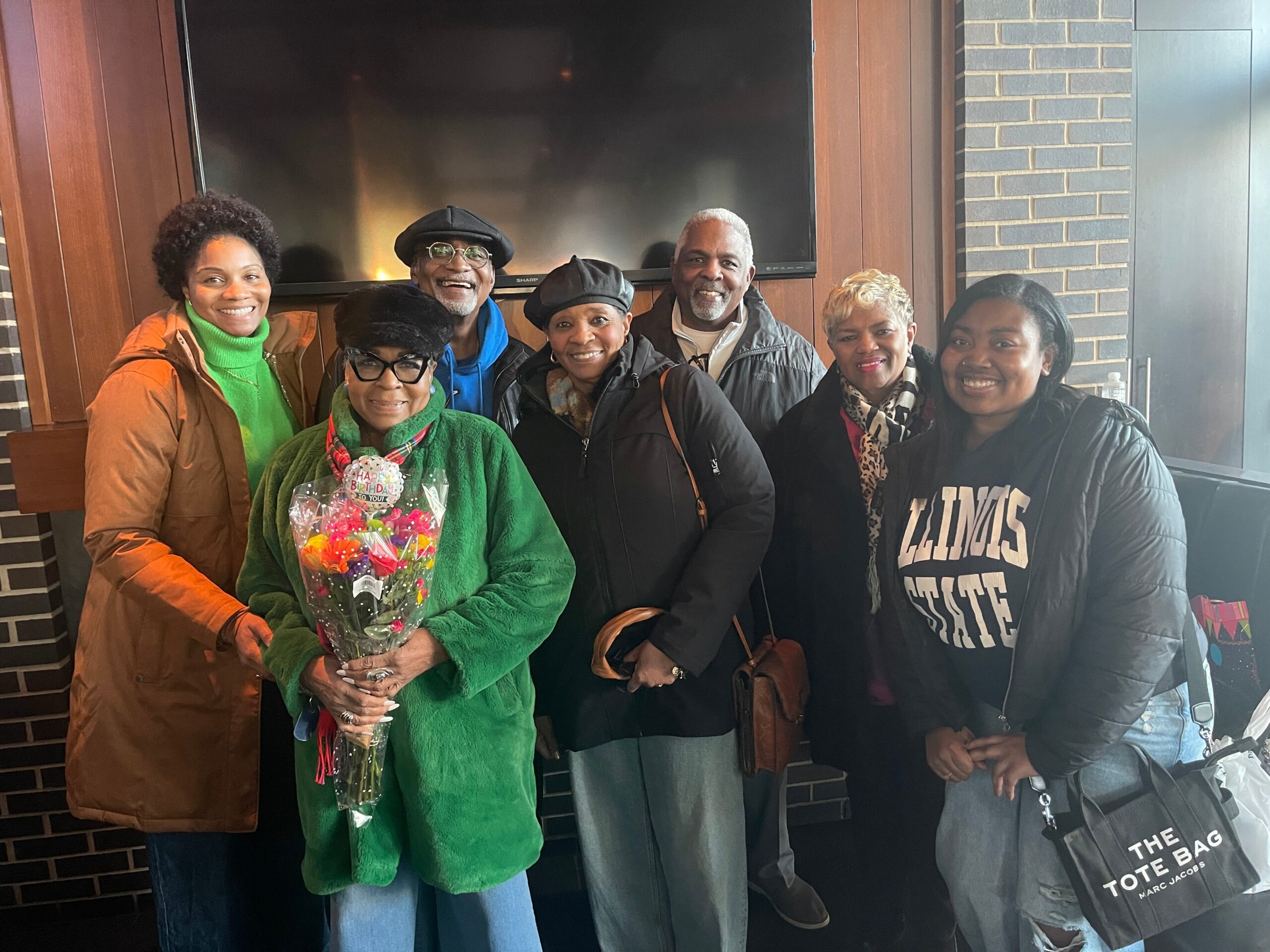Six adults smiling for a group photo indoors near a brick wall and TV; woman in green holds a bouquet.