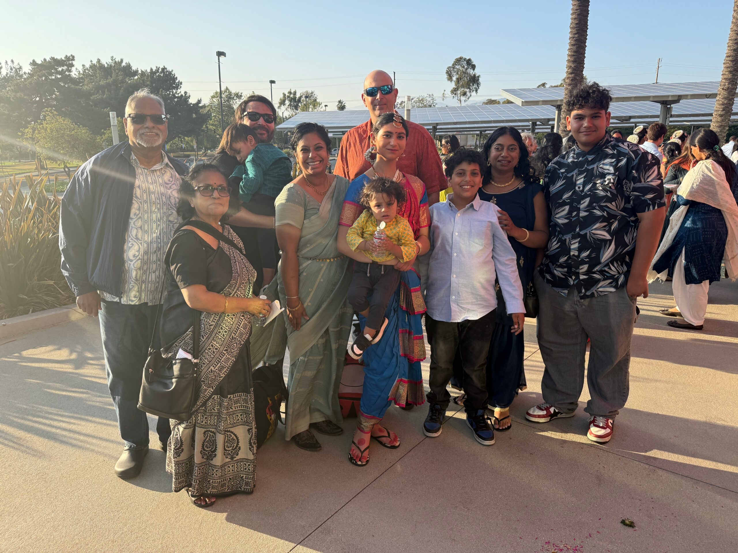 Group of Indian people posing together outdoors on a sunny day, wearing traditional sarees and casual clothing, smiling.